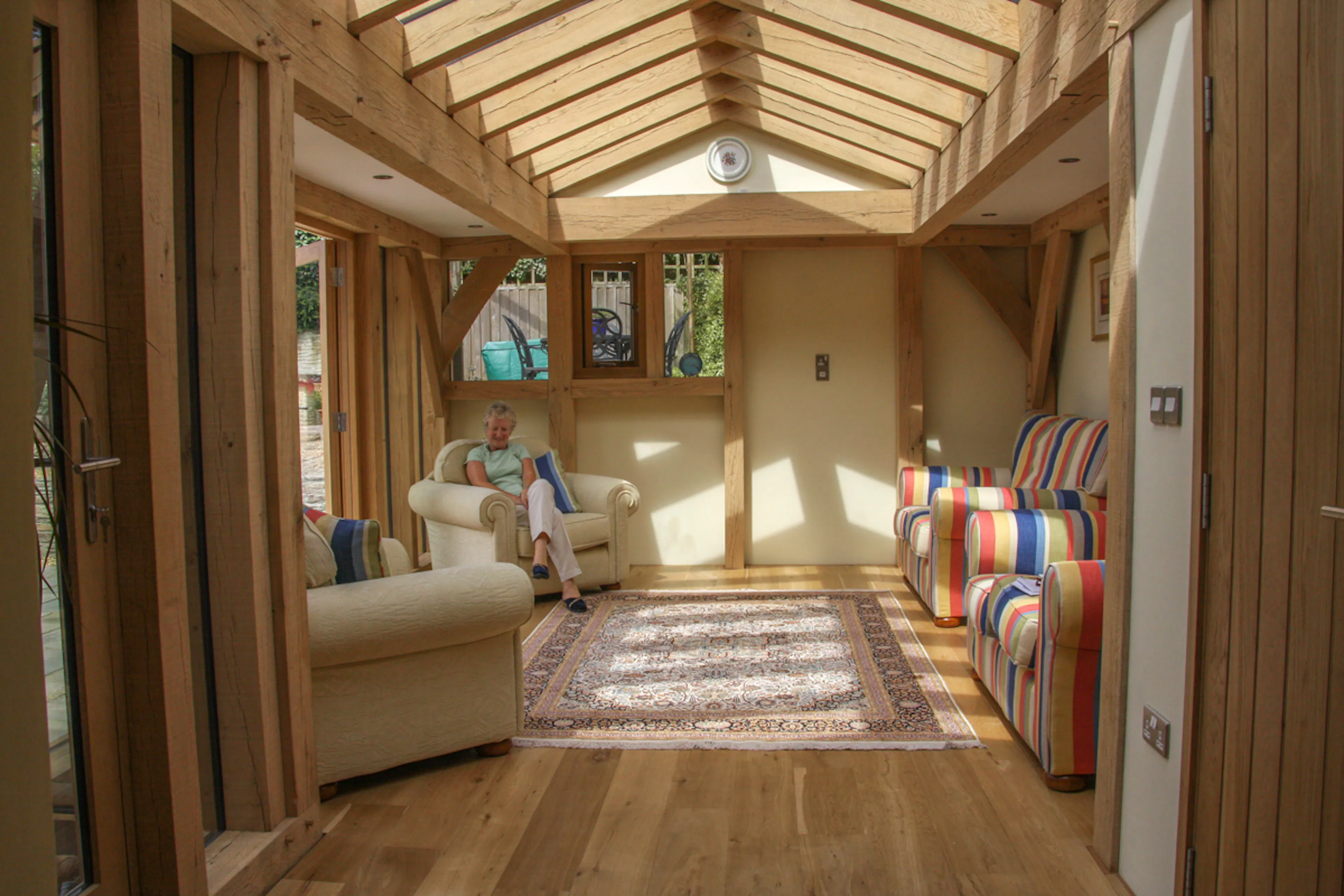 An oak framed sun room or garden room extension with sunlight entering through glazed rooflights and windows, and furnished with sofas and armchairs.