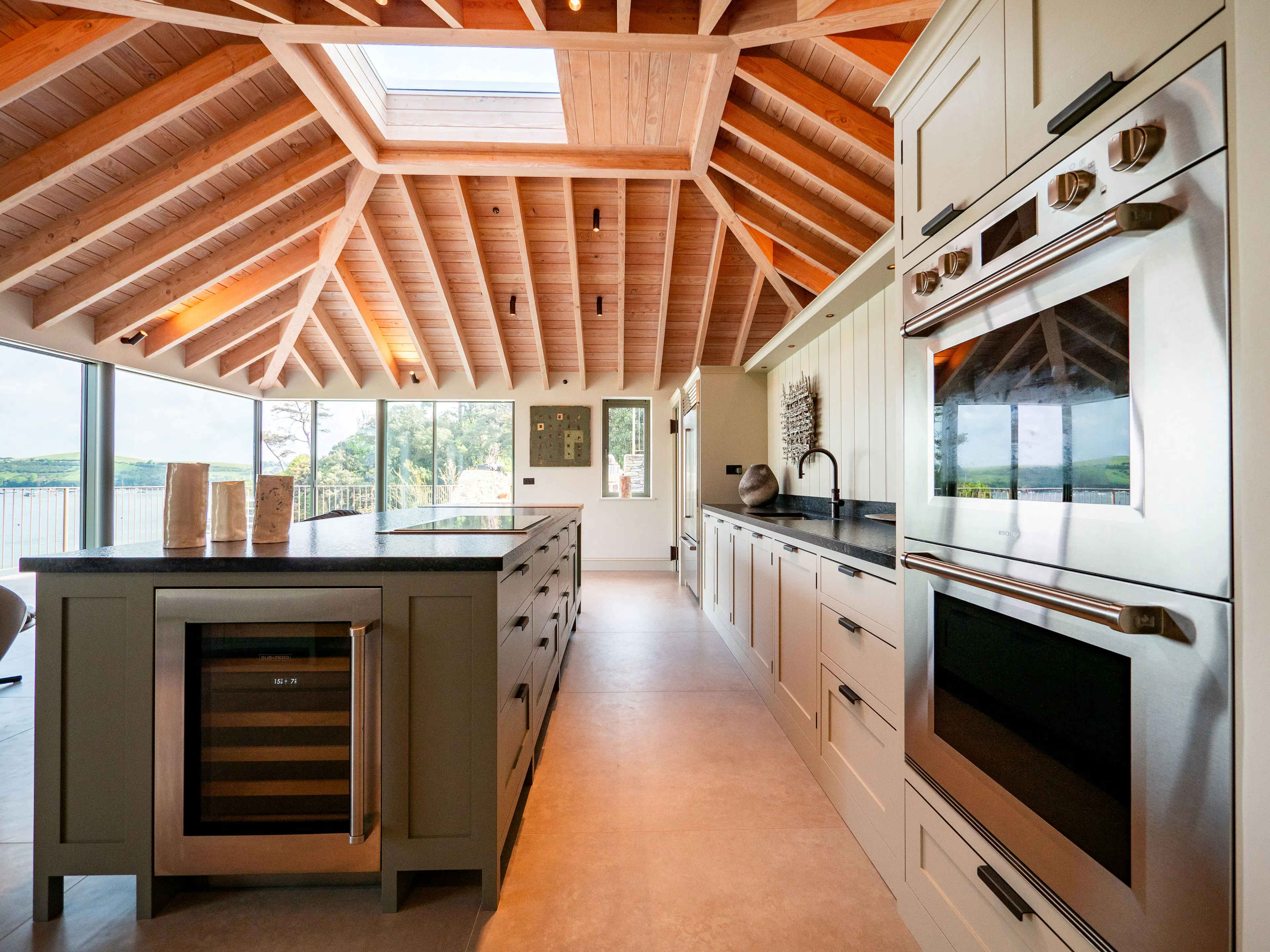 A contemporary Douglas fir glulam framed kitchen extension with a large kitchen island and modern armchairs by glazed sliding doors