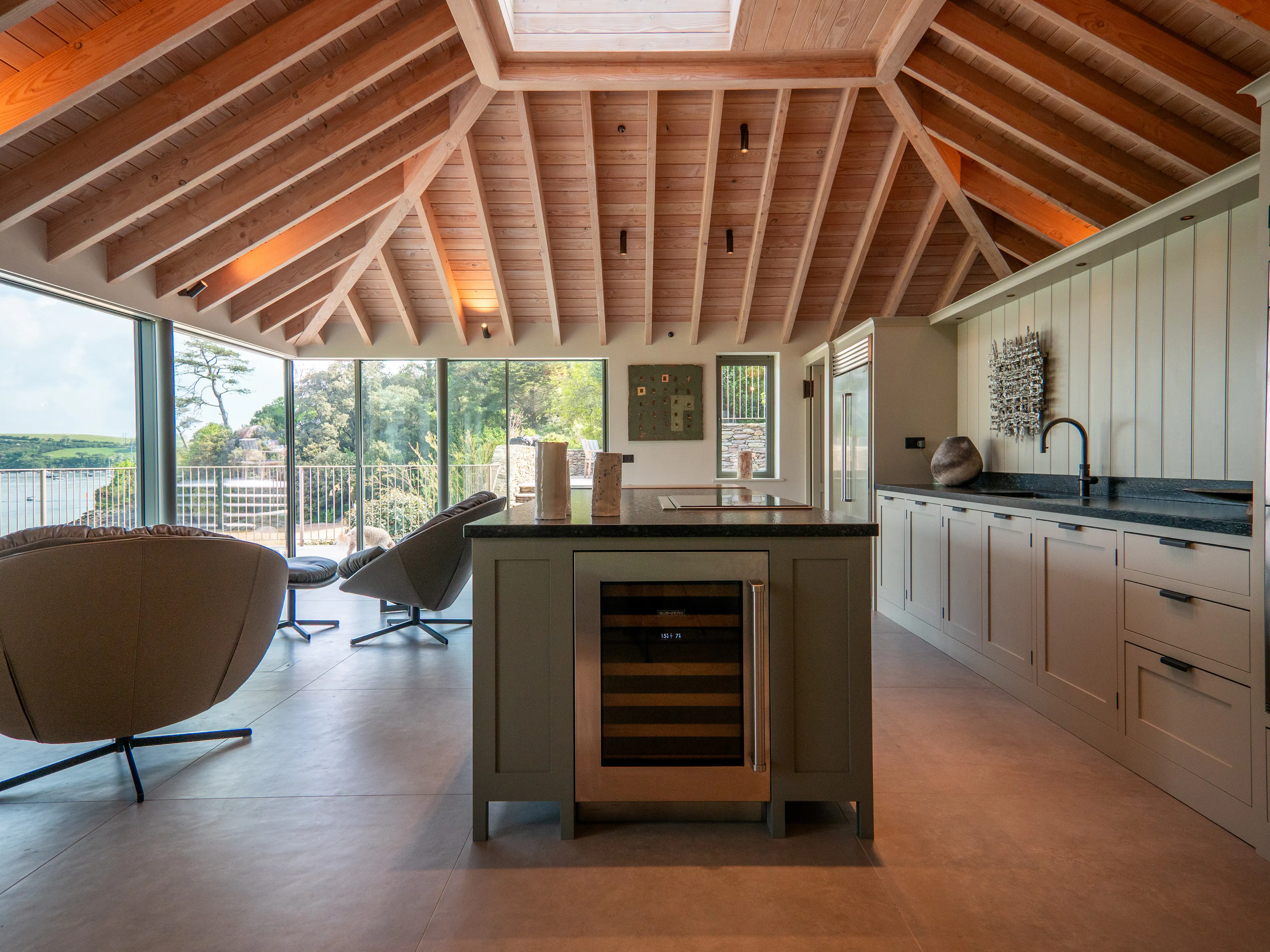 A contemporary Douglas fir glulam framed kitchen extension with a large kitchen island and modern armchairs by glazed sliding doors