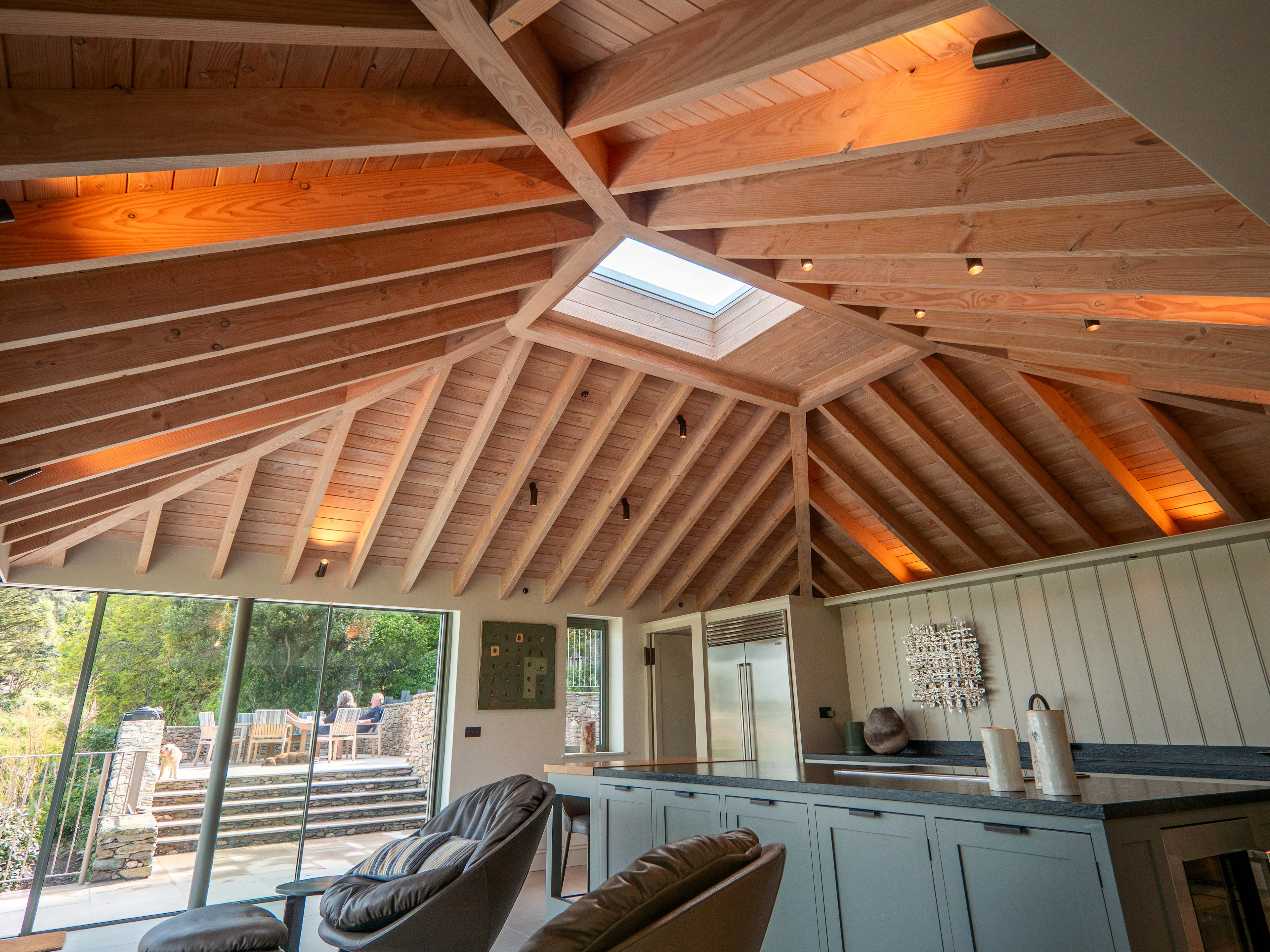 A contemporary Douglas fir glulam framed kitchen extension with a large kitchen island and modern armchairs by glazed sliding doors