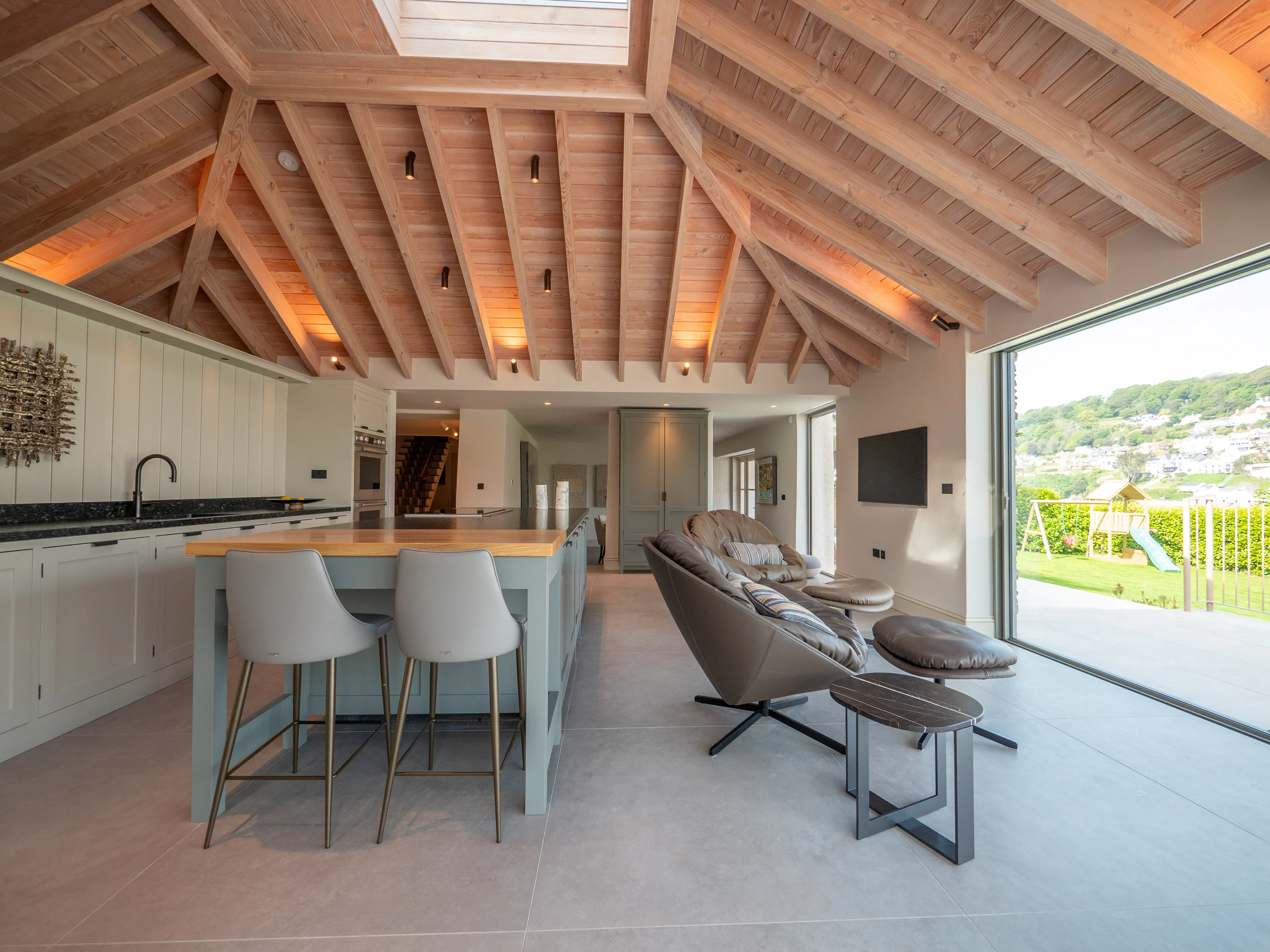 A contemporary Douglas fir glulam framed kitchen extension with a large kitchen island and modern armchairs by glazed sliding doors