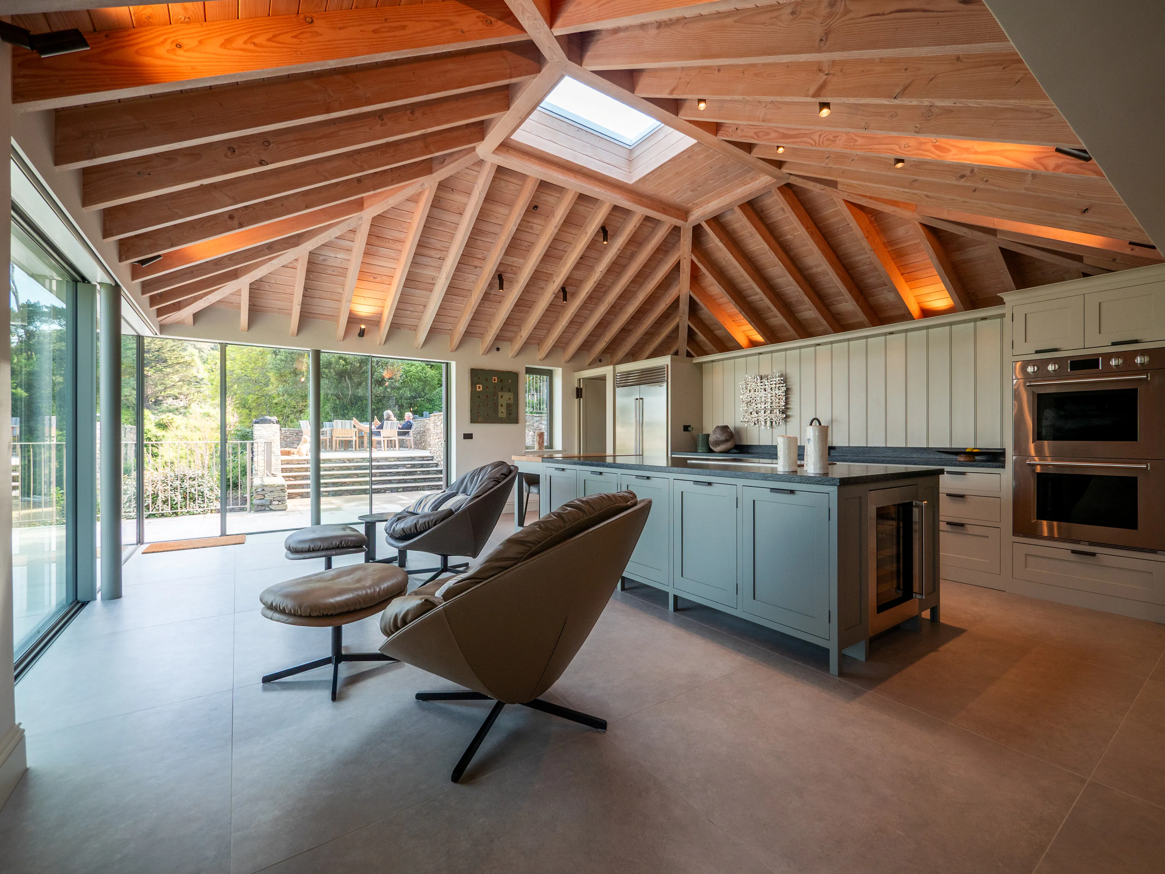 A contemporary Douglas fir glulam framed kitchen extension with a large kitchen island and modern armchairs by glazed sliding doors