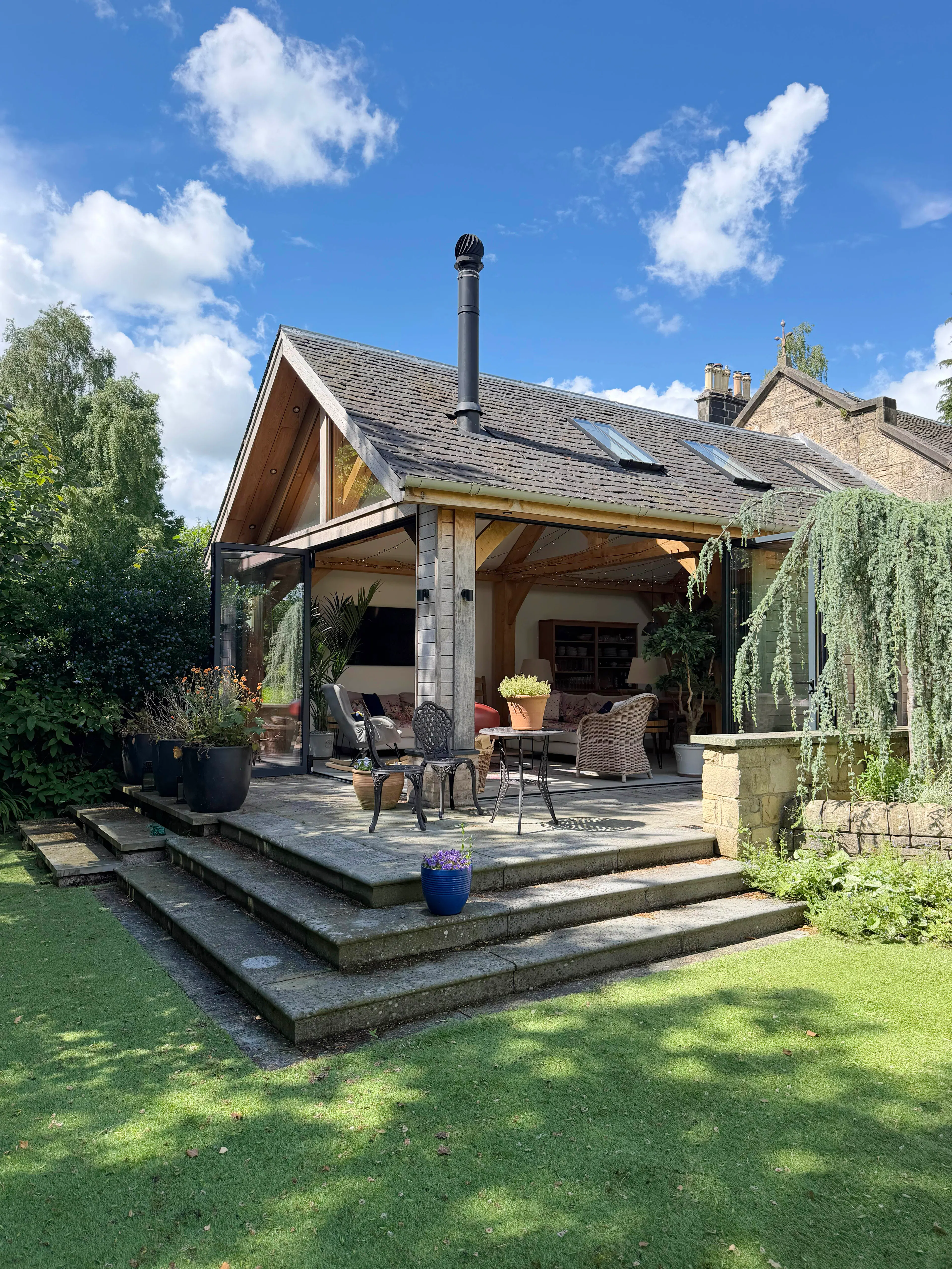 Garden room with oak frame construction, bifold doors, timber cladding, and a slate roof, featuring landscaped garden planting, patio paving, and steps leading to the entrance