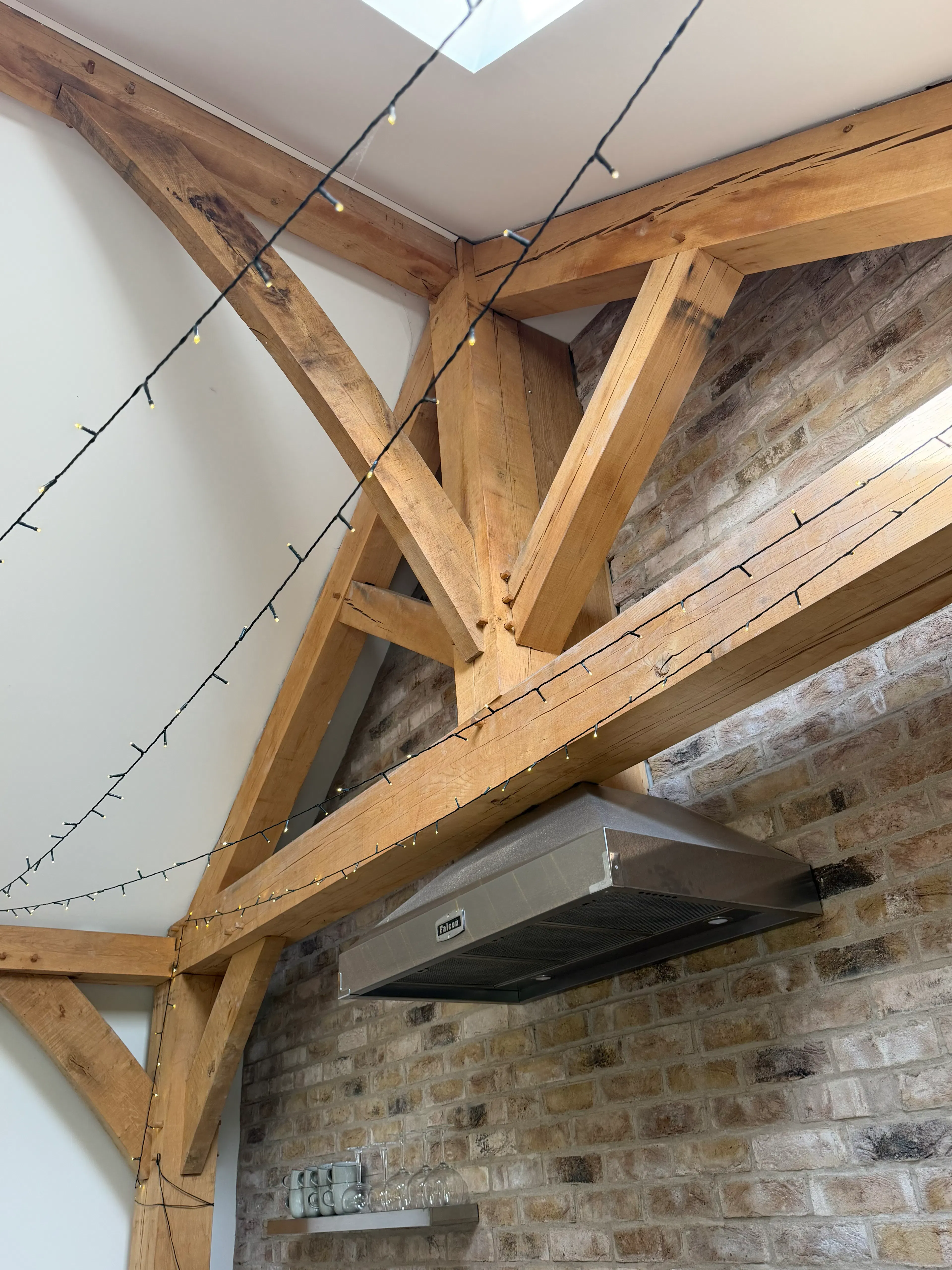 Open-plan garden room interior with exposed oak frame construction and a king post truss, featuring a kitchen area and structural wall brace within a bright living space