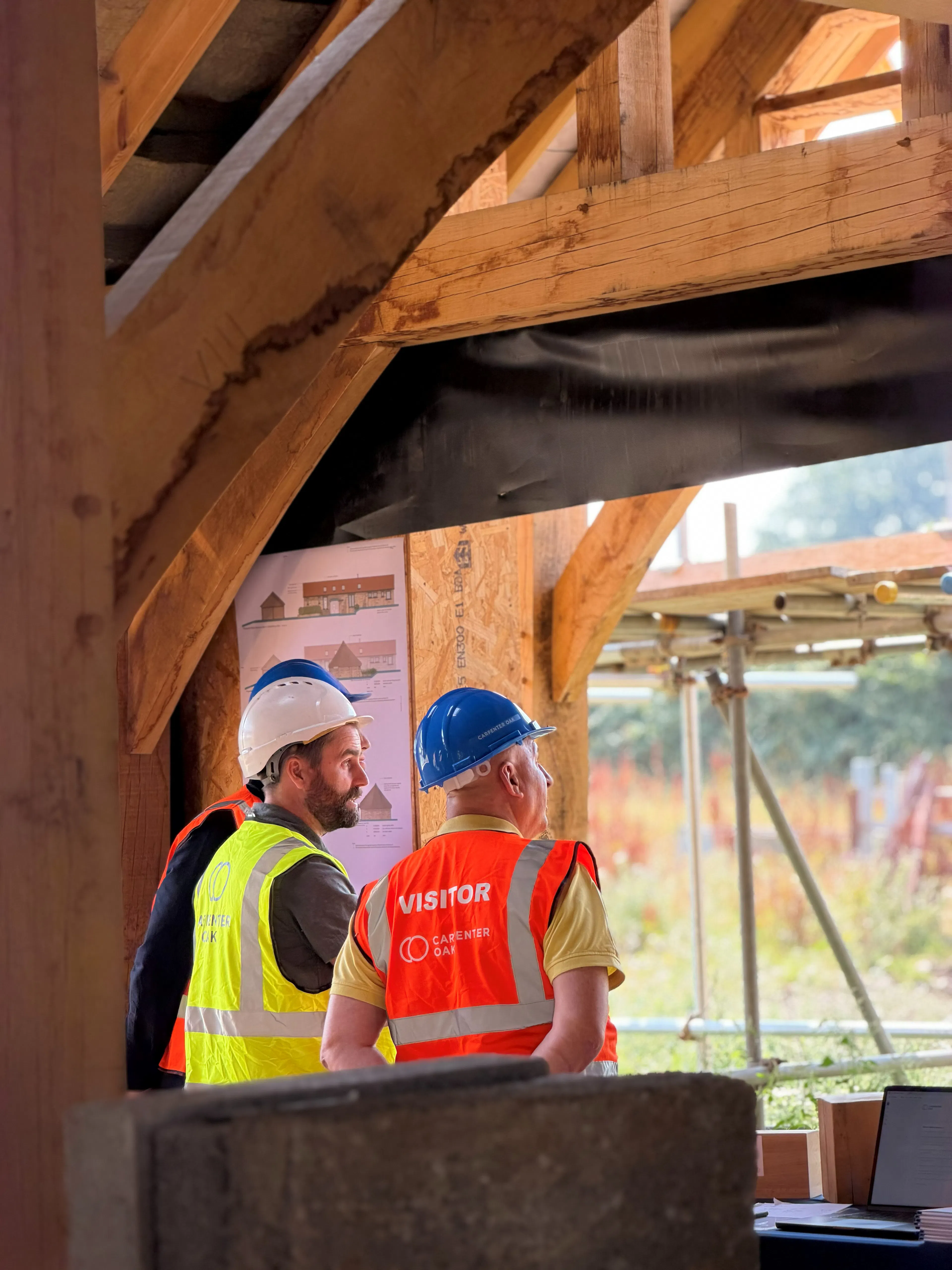 An event on a construction site where attendees in high vis jackets and blue hard hats are shown around an oak frame house mid-build