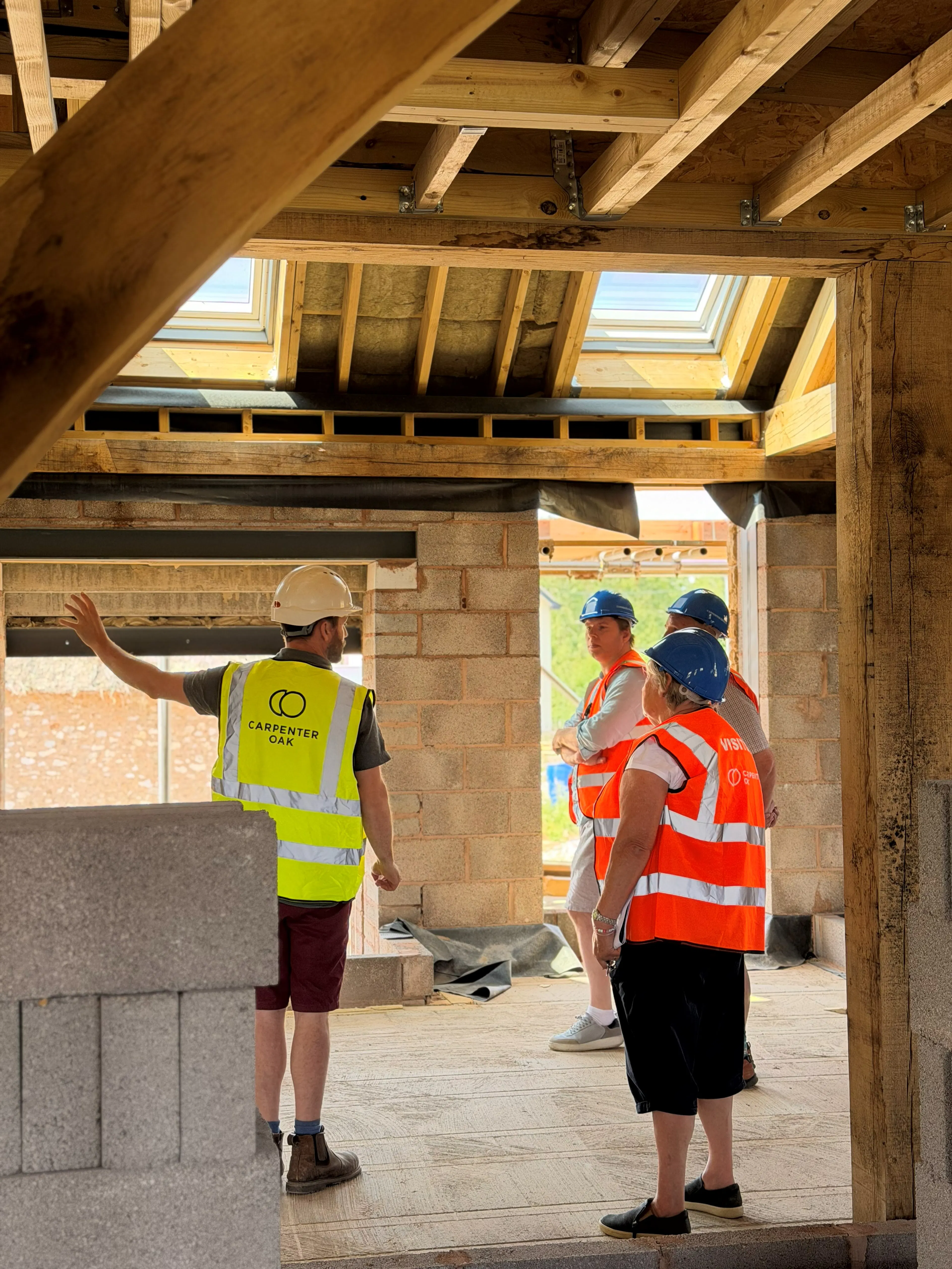 An event on a construction site where attendees in high vis jackets and blue hard hats are shown around an oak frame house mid-build