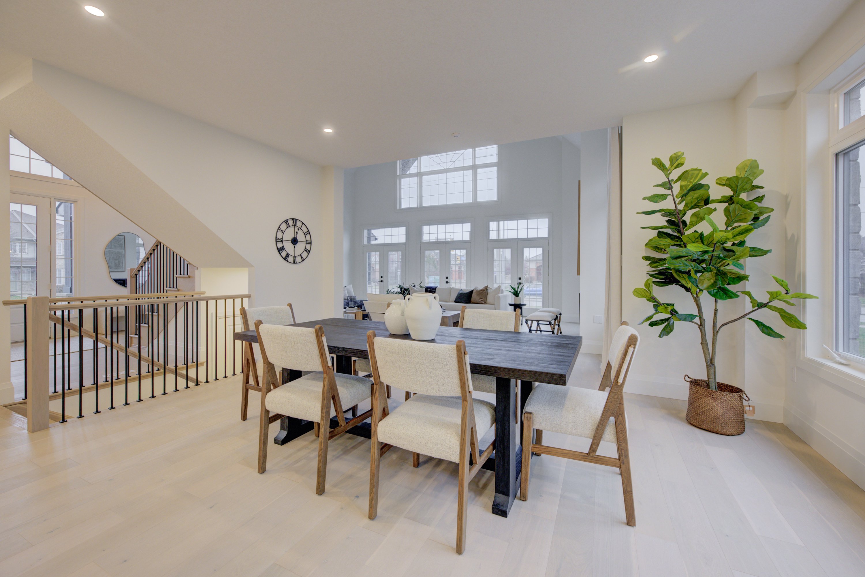 Bright dining area with a dark wood table, cream chairs, and large windows, adjacent to an open staircase