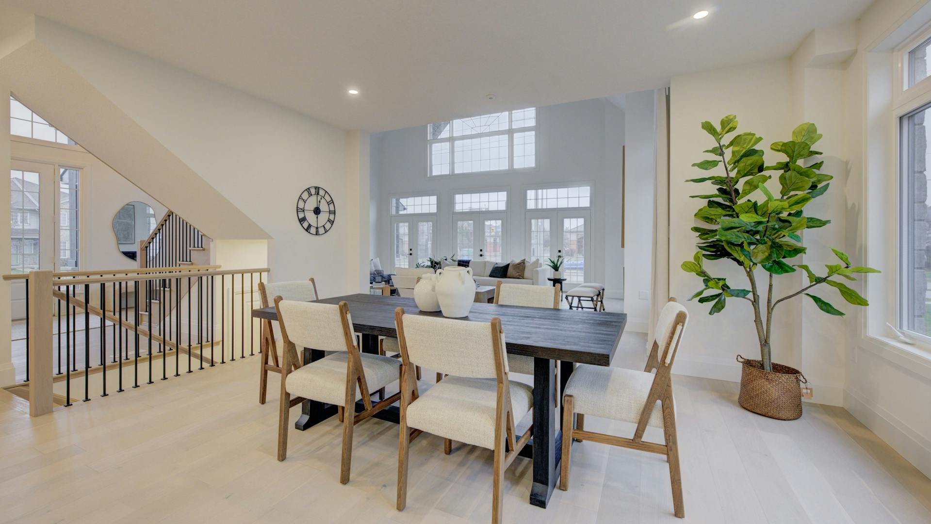 Bright dining area with a dark wood table, cream chairs, and large windows, adjacent to an open staircase