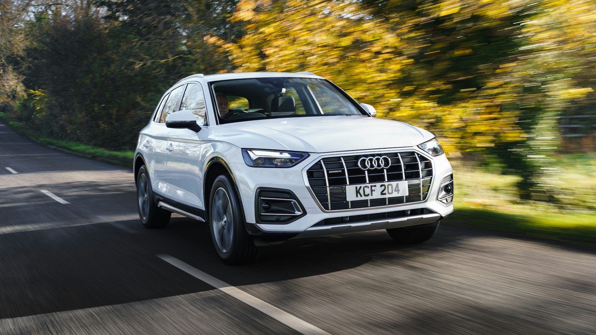 White 2020 Audi Q5 driving in the countryside beside autumn trees, front quarter view.