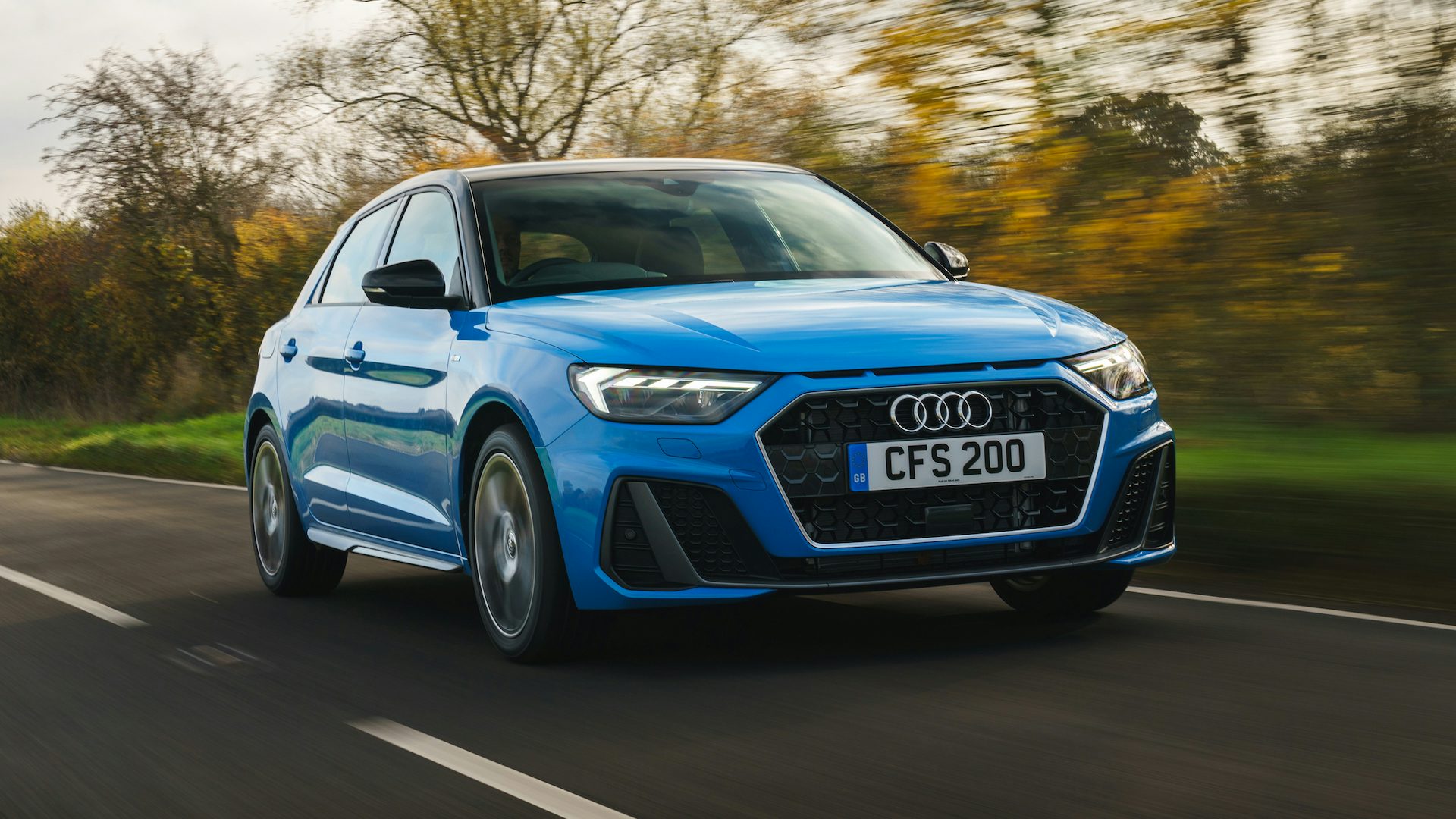 Blue 2018 Audi A1 driving in the countryside with a low sun behind winter trees, front quarter view.