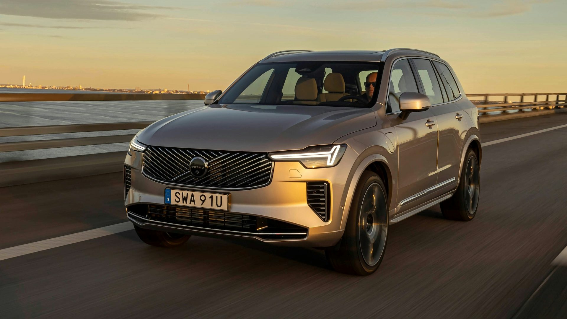 Silver 2025 Volvo XC90 driving next to the ocean at sunset, front quarter angle.
