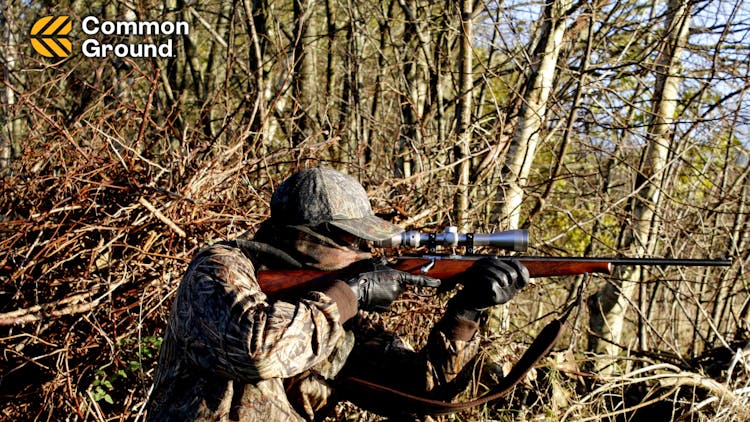 A man wearing hunting gear aiming a rifle in a forest.
