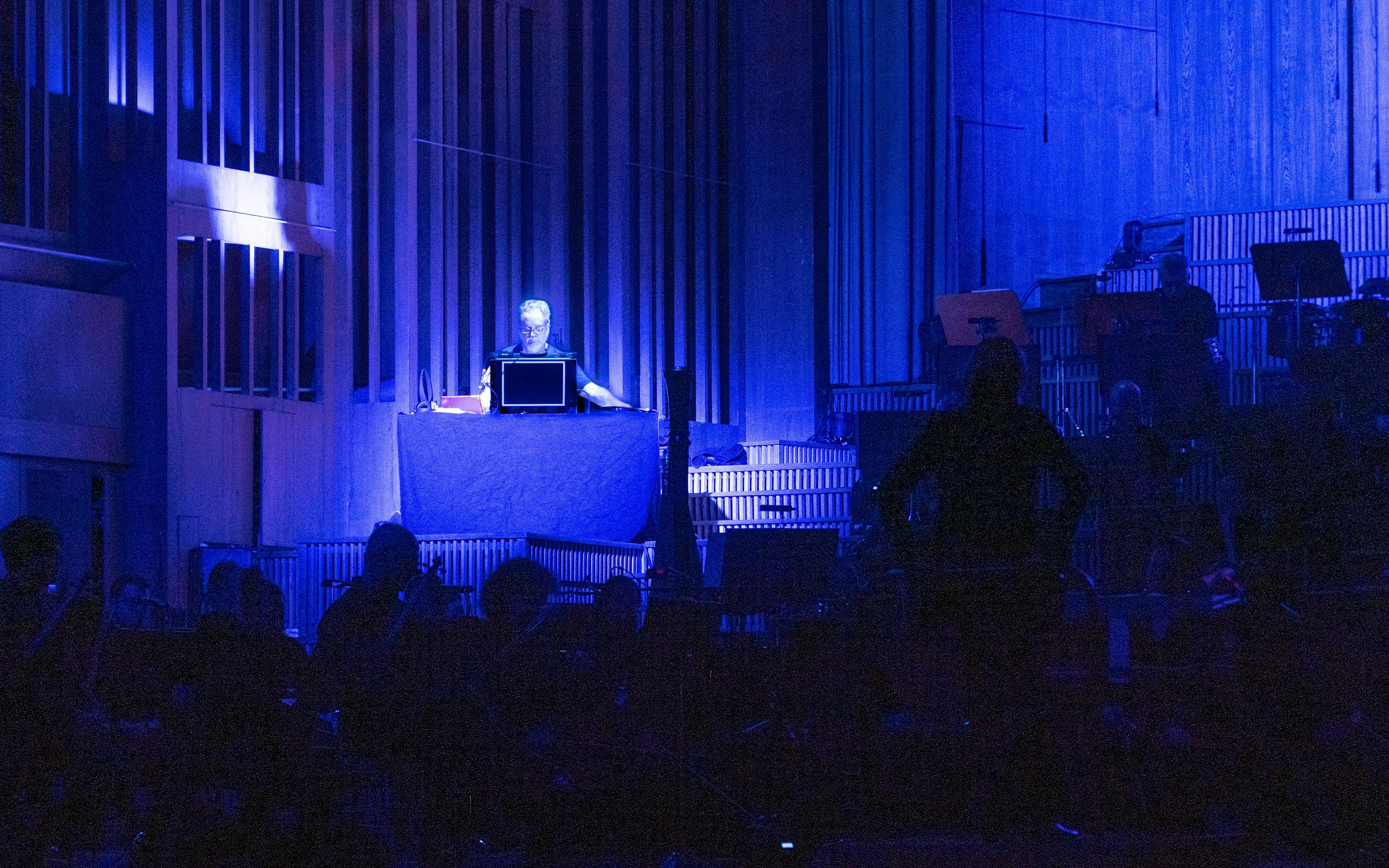 Man on stage plays modular synthesiser alongside an orchestra and under a spotlight 