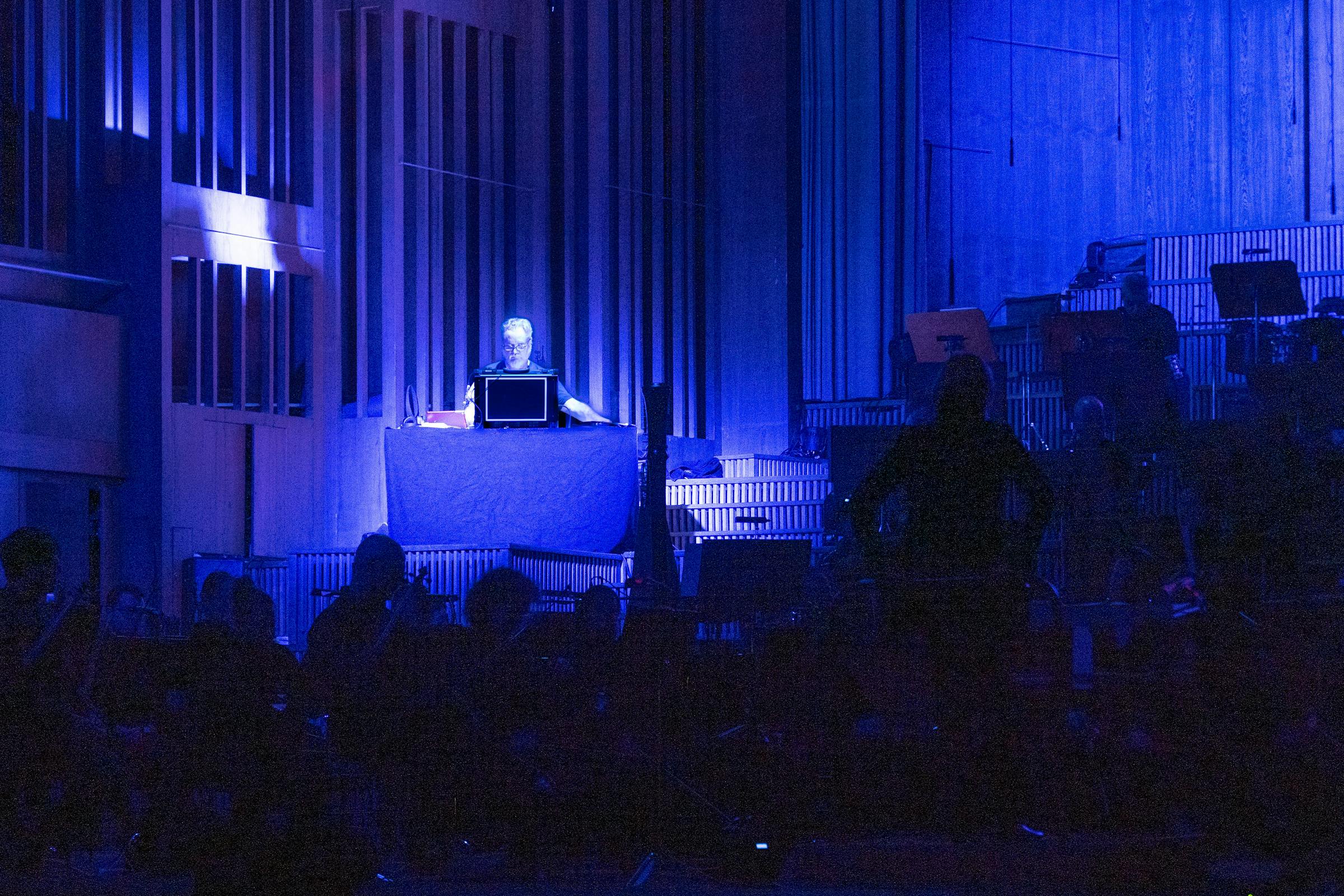 Man on stage plays modular synthesiser alongside an orchestra and under a spotlight 