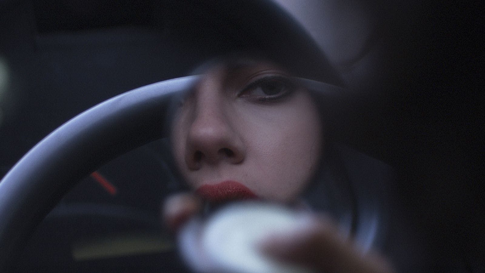 A woman looks at herself in a pocket mirror, behind the mirror is a steering wheel