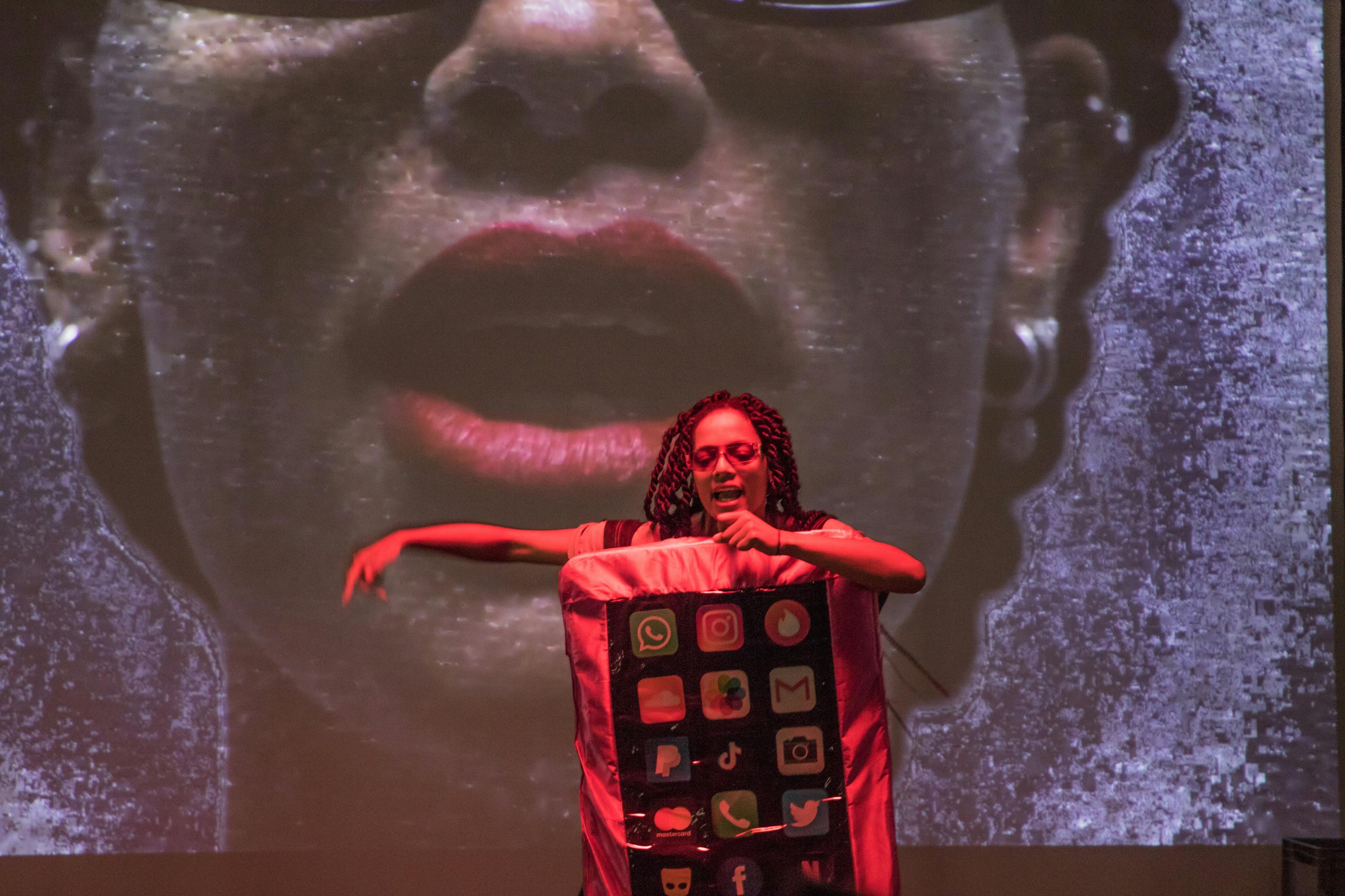 Shanice Trustfull's monologue. She wears a suit like an iphone, showing different apps on her front. Behind is her portrait on a large screen