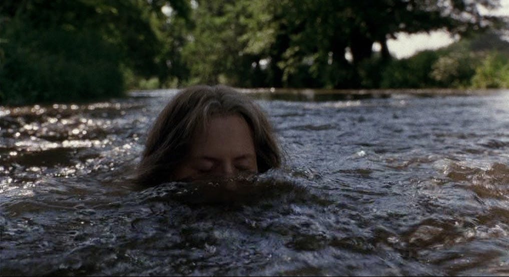 A woman submerges her whole body into a running river, only the top of her head and closed eyes are above the waterline