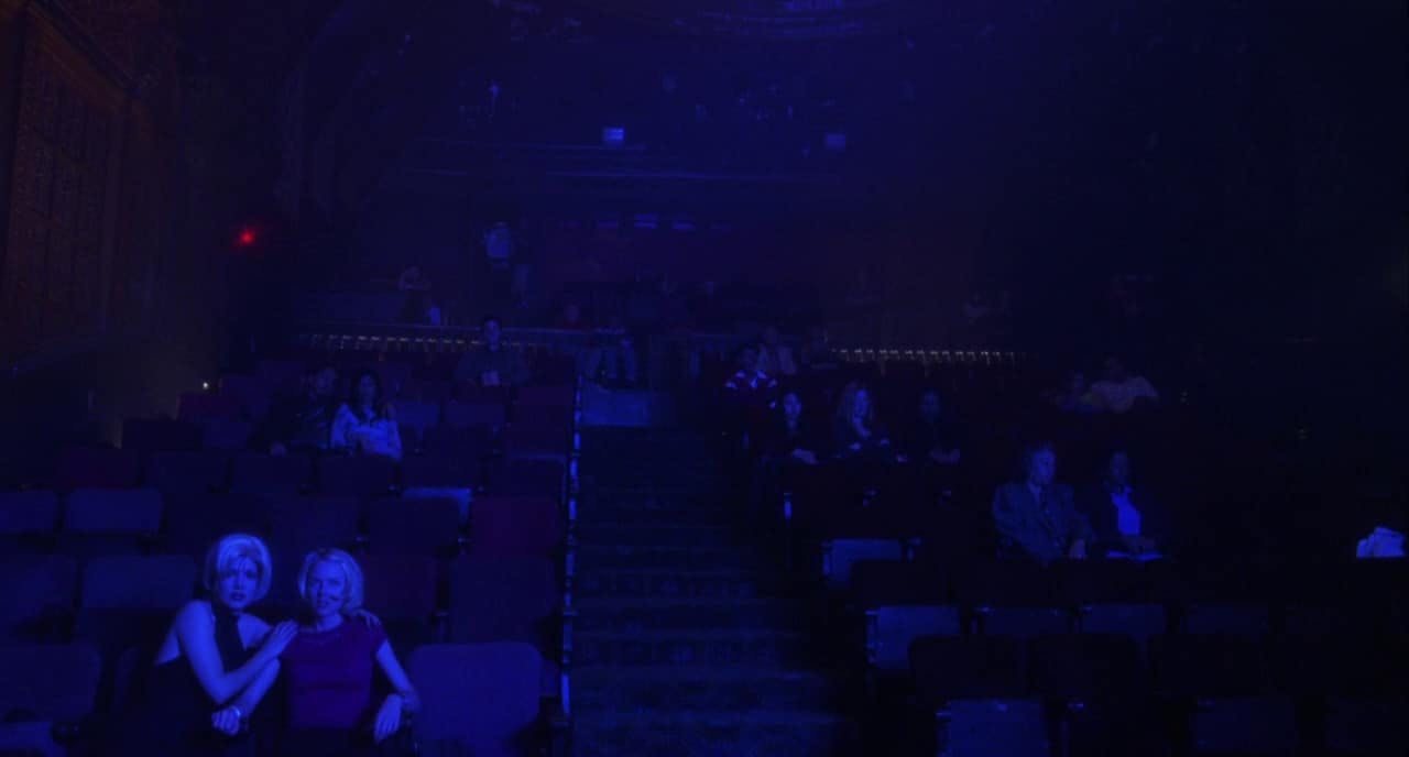 A dimly lit theater filled with people seated in rows. The front two seats are taken by two protagonists