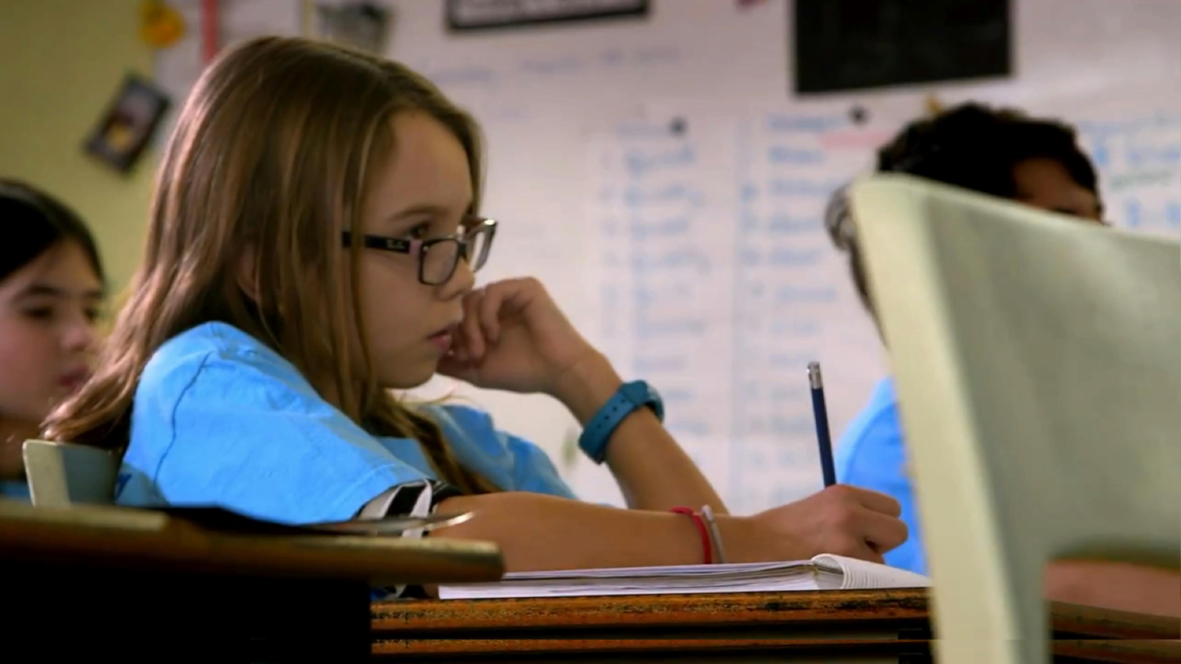 A girl in a blue shirt writes in a notebook at her desk.