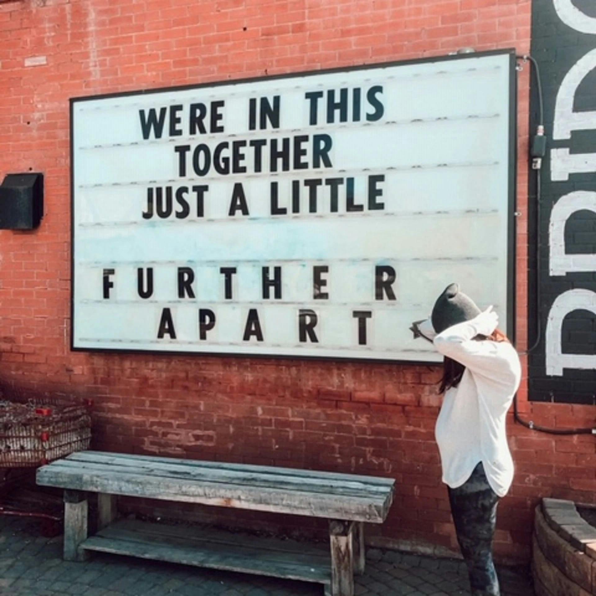 Woman standing in front of sign that says 'We're in this together, just a little further apart'