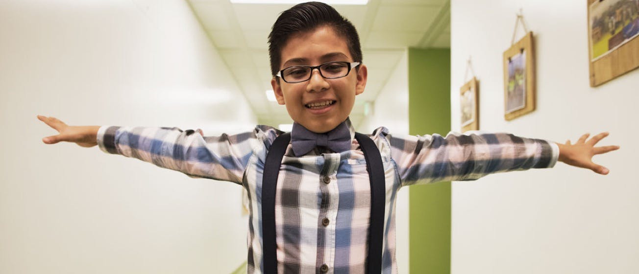 A boy with glasses and plaid button up shirt is raising his hands looking happy
