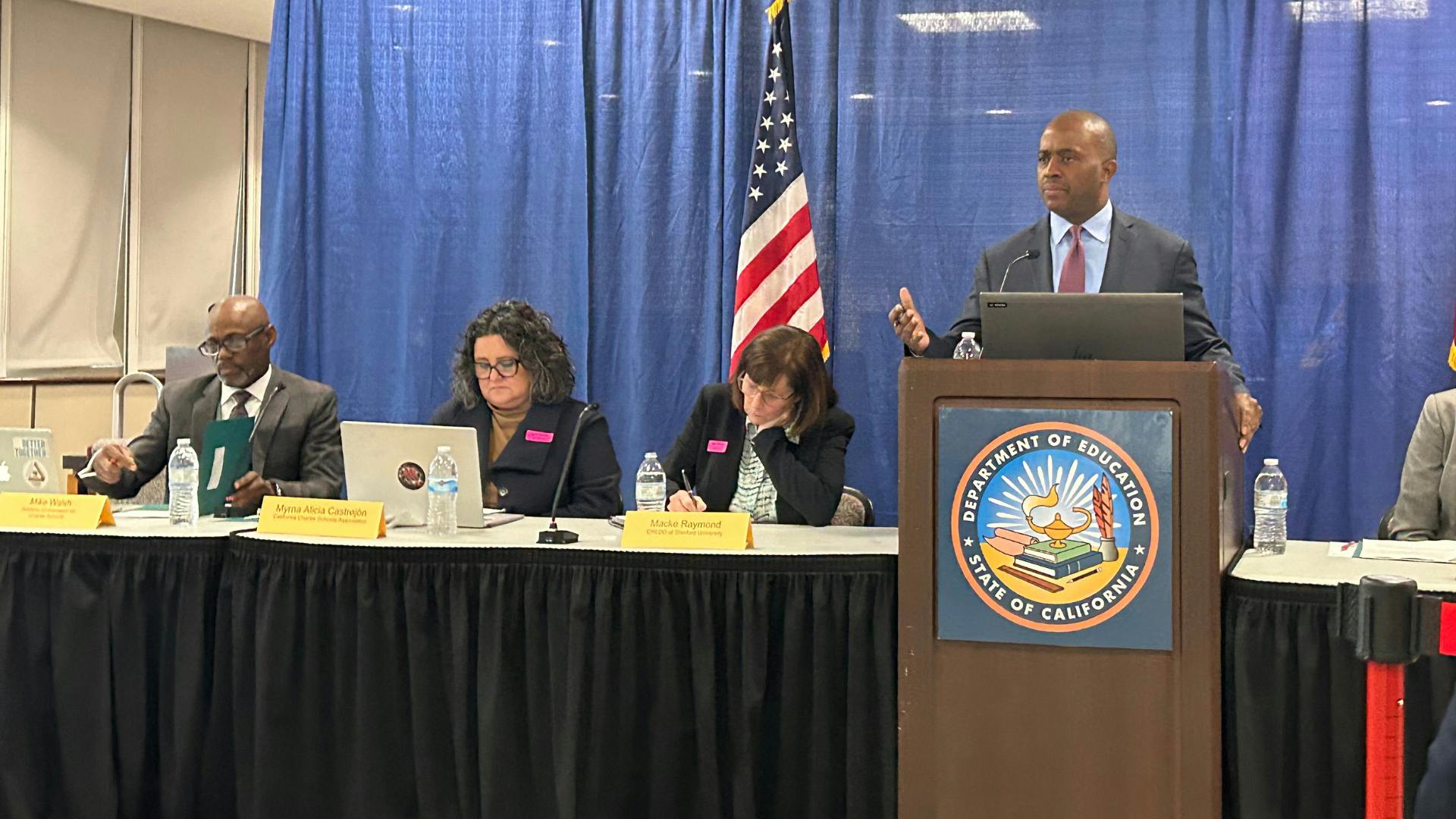 Photo of the State Superintendent of Public Instruction, Tony Thurmond, leading a panel on funding. Three people are sitting down with nameplates in front of them and one of them is Myrna Castrejón, President and Chief Executive Officer California Charter Schools Association.