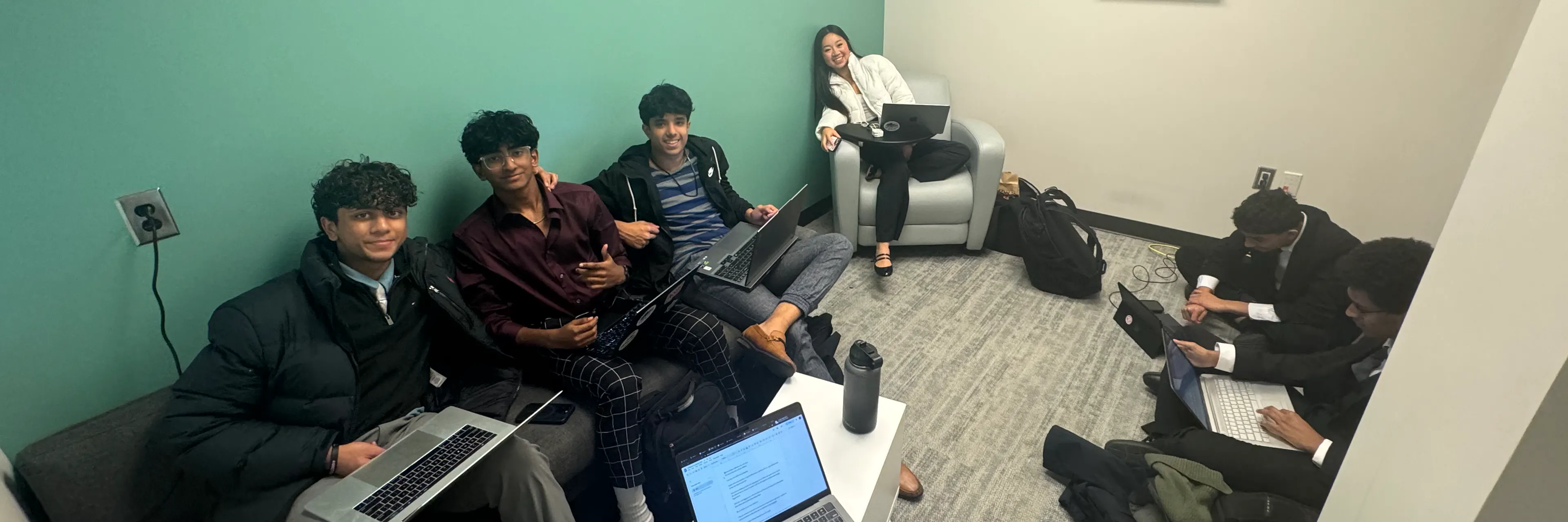 A group of teen students with laptops sit in a room and smile at the camera