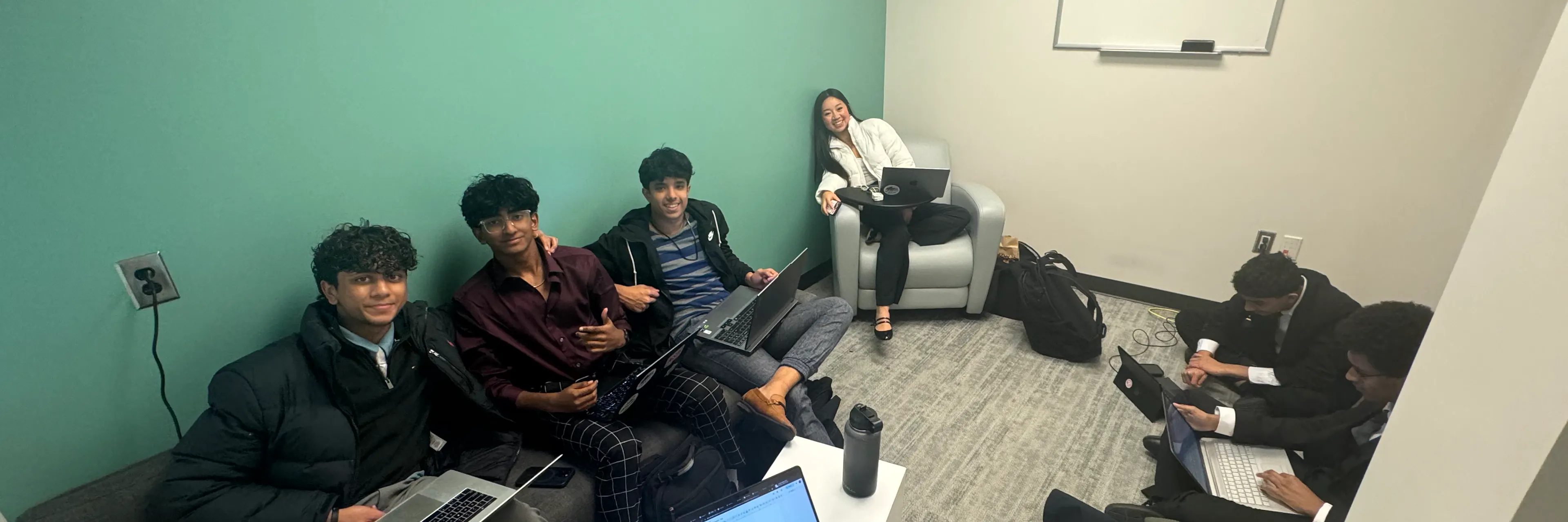 A group of teen students with laptops sit in a room and smile at the camera