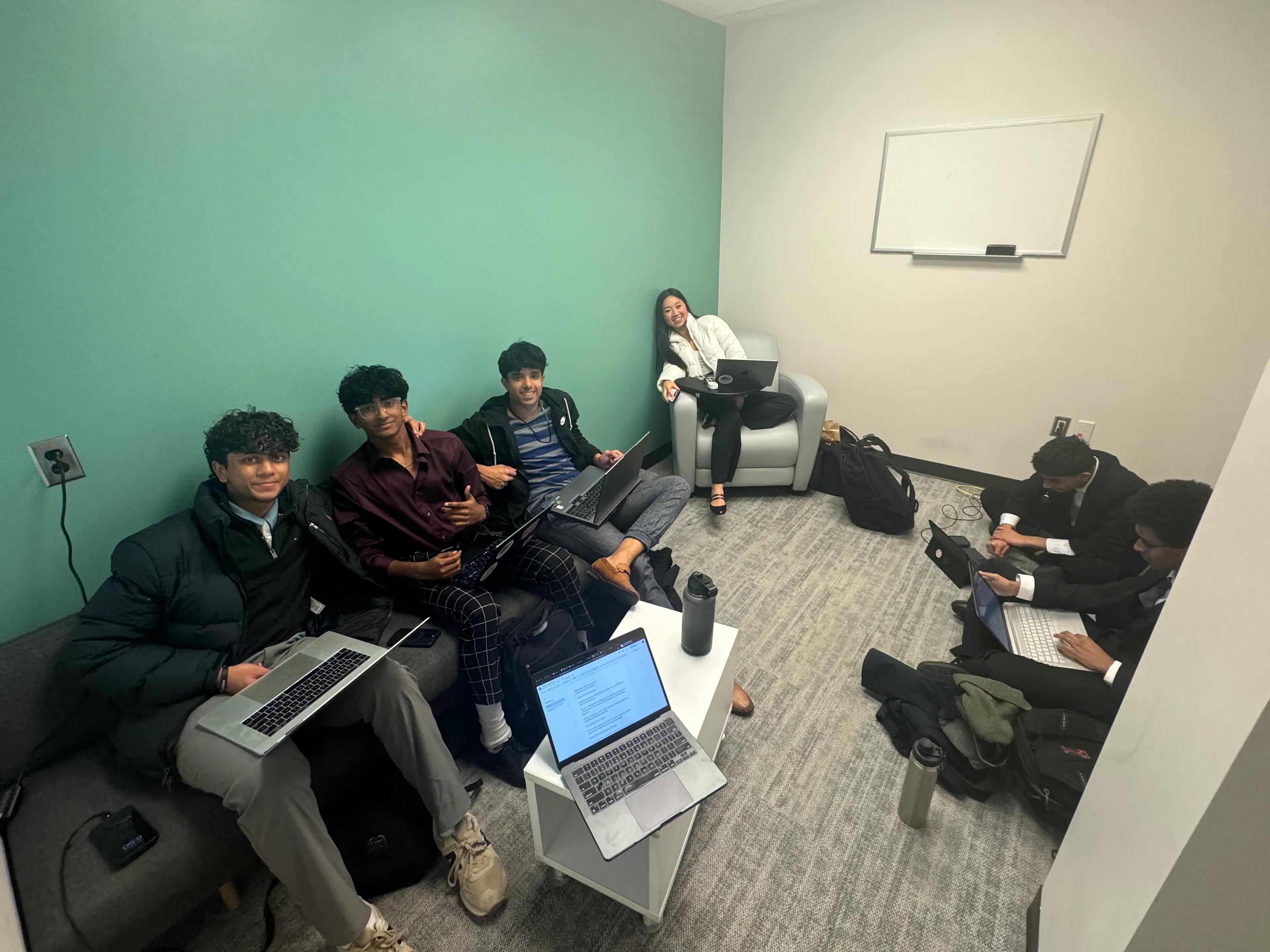 A group of teen students with laptops sit in a room and smile at the camera