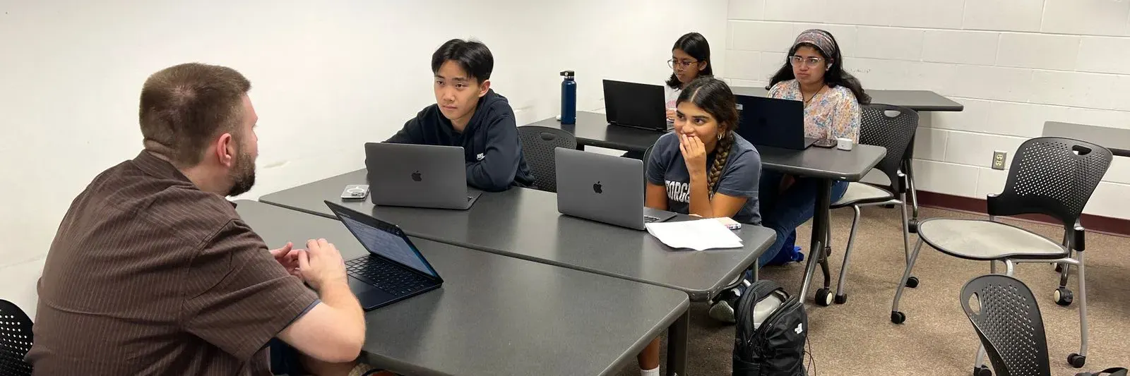 Four students with laptops sit at tables in front of an instructor