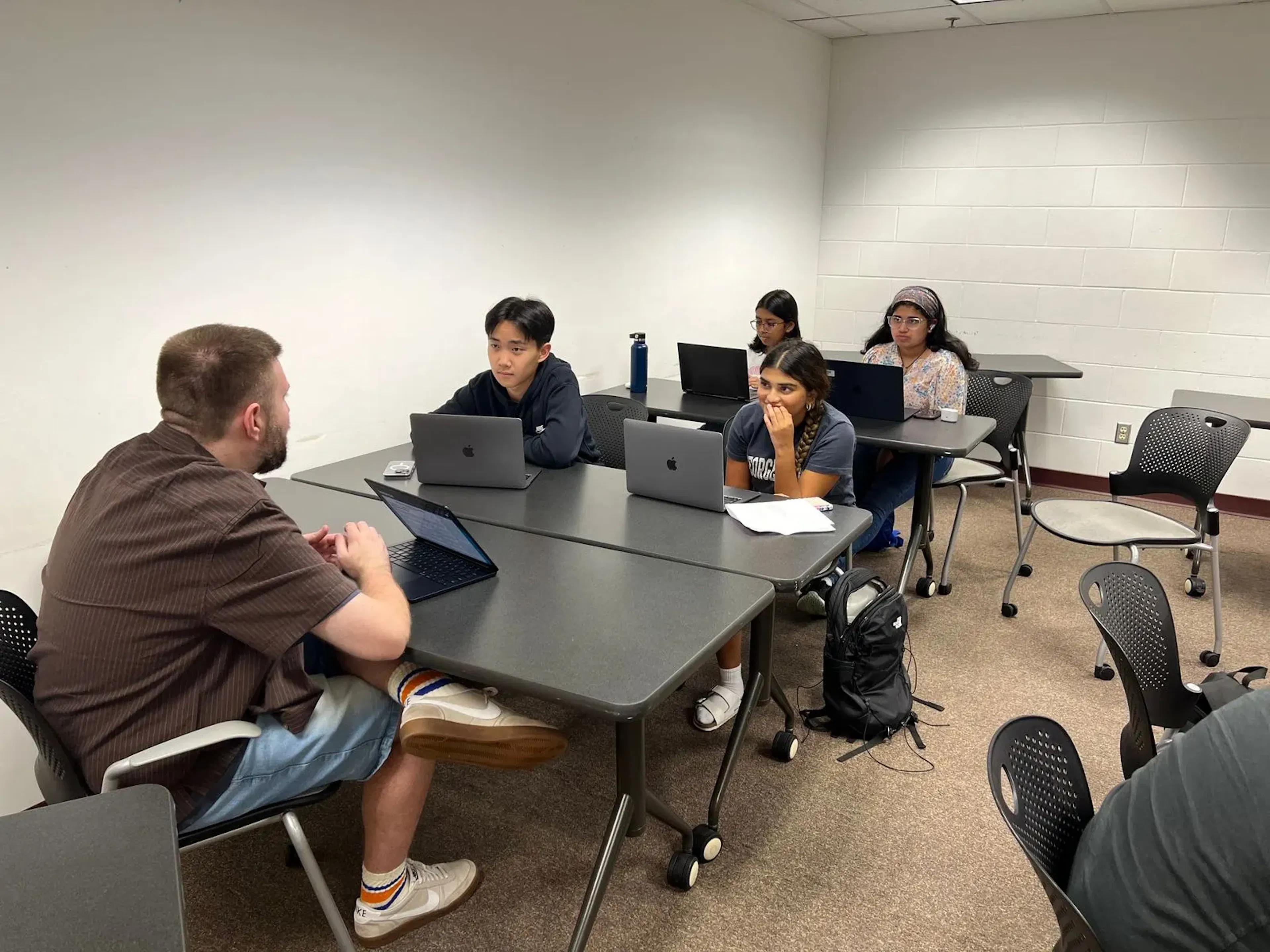 Four students with laptops sit at tables in front of an instructor