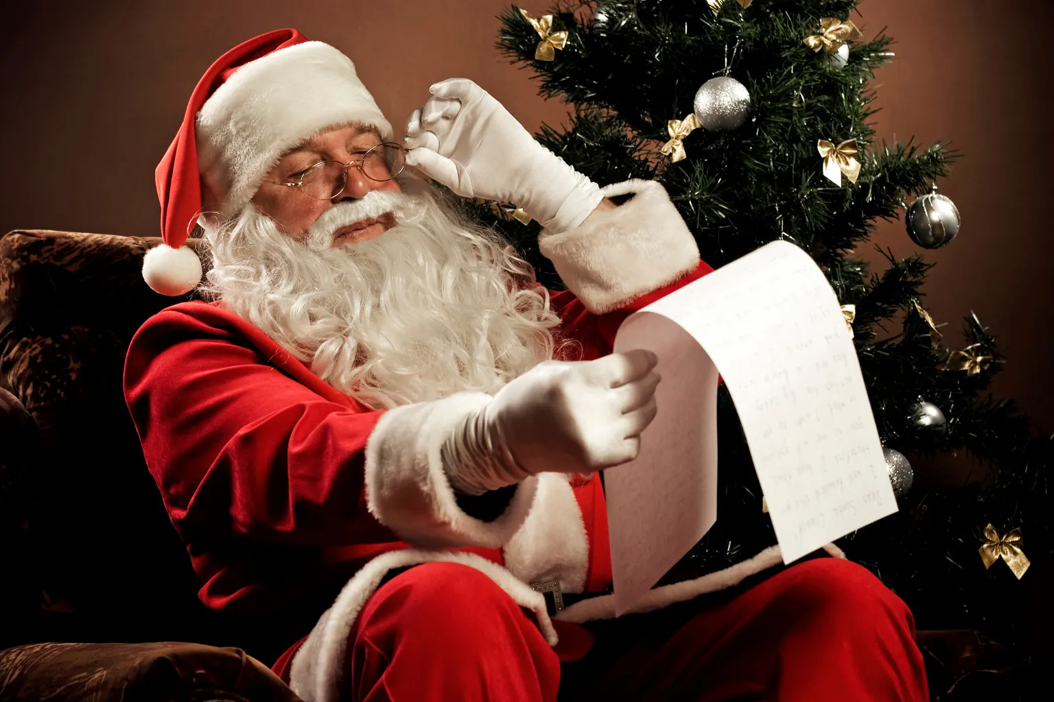 Santa reading a child’s Christmas letter beside a decorated tre