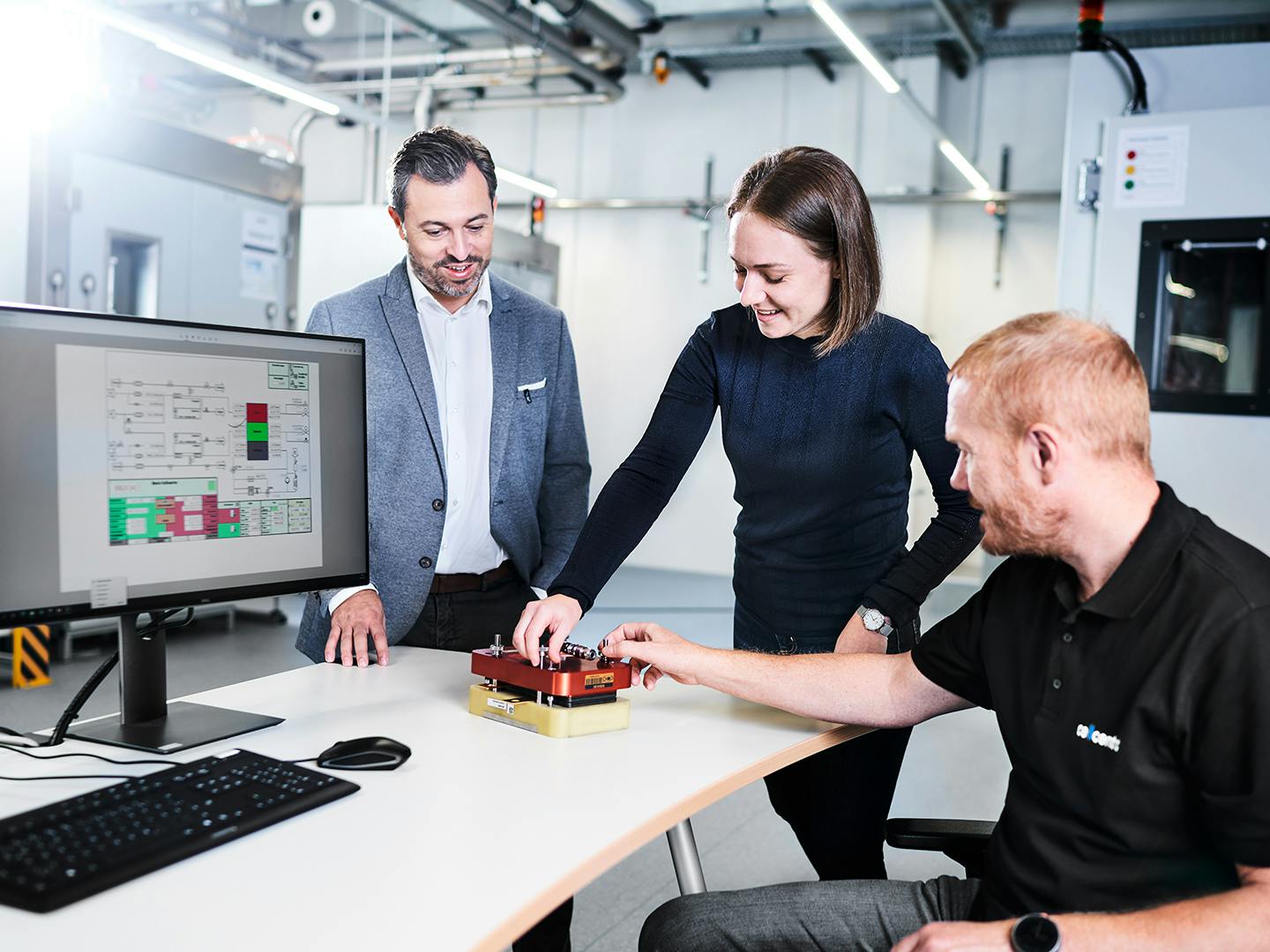 Three employees test a component at a table, while a monitor displays the manufacturing diagram in the production hall.