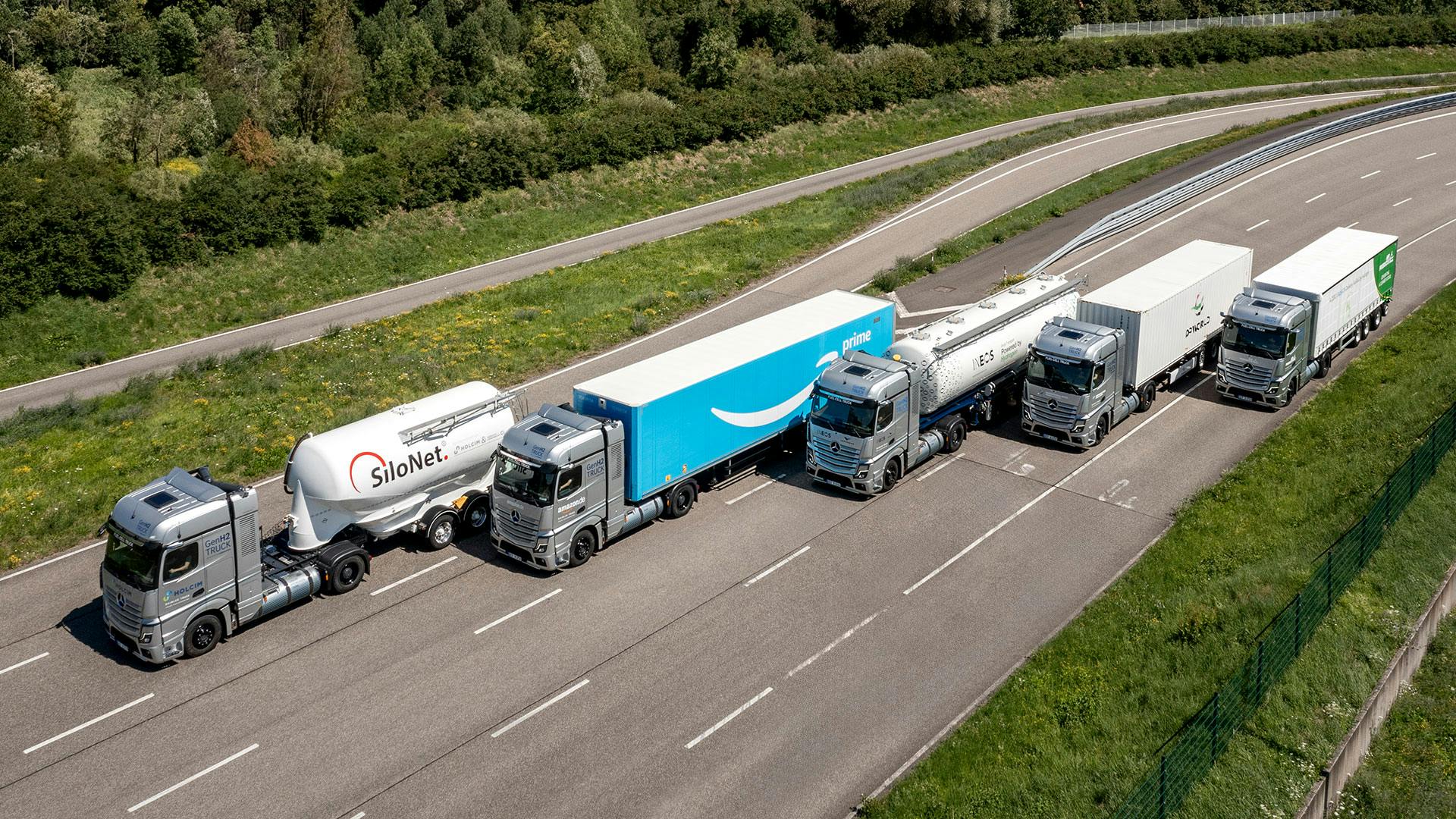 Fünf moderne LKWs fahren im Kolonne-Testbetrieb auf einer Asphaltstrecke neben grüner Böschung.