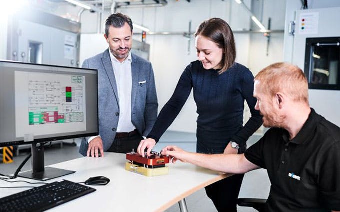 Drei Mitarbeitende testen ein Bauteil am Tisch, Monitor zeigt Fertigungsschema in Produktionshalle.