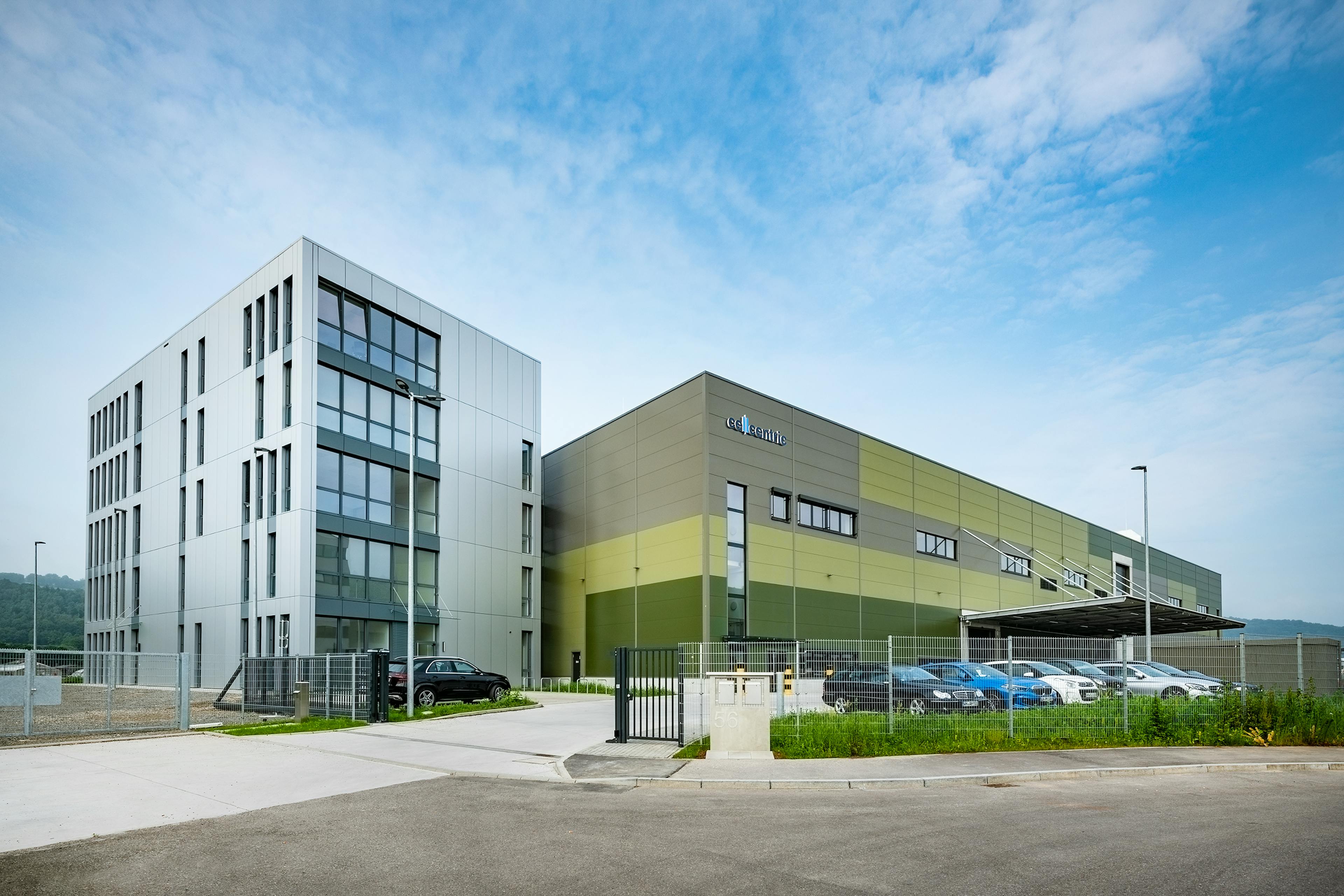 Modern company building with office wing and production hall behind a fenced parking area.