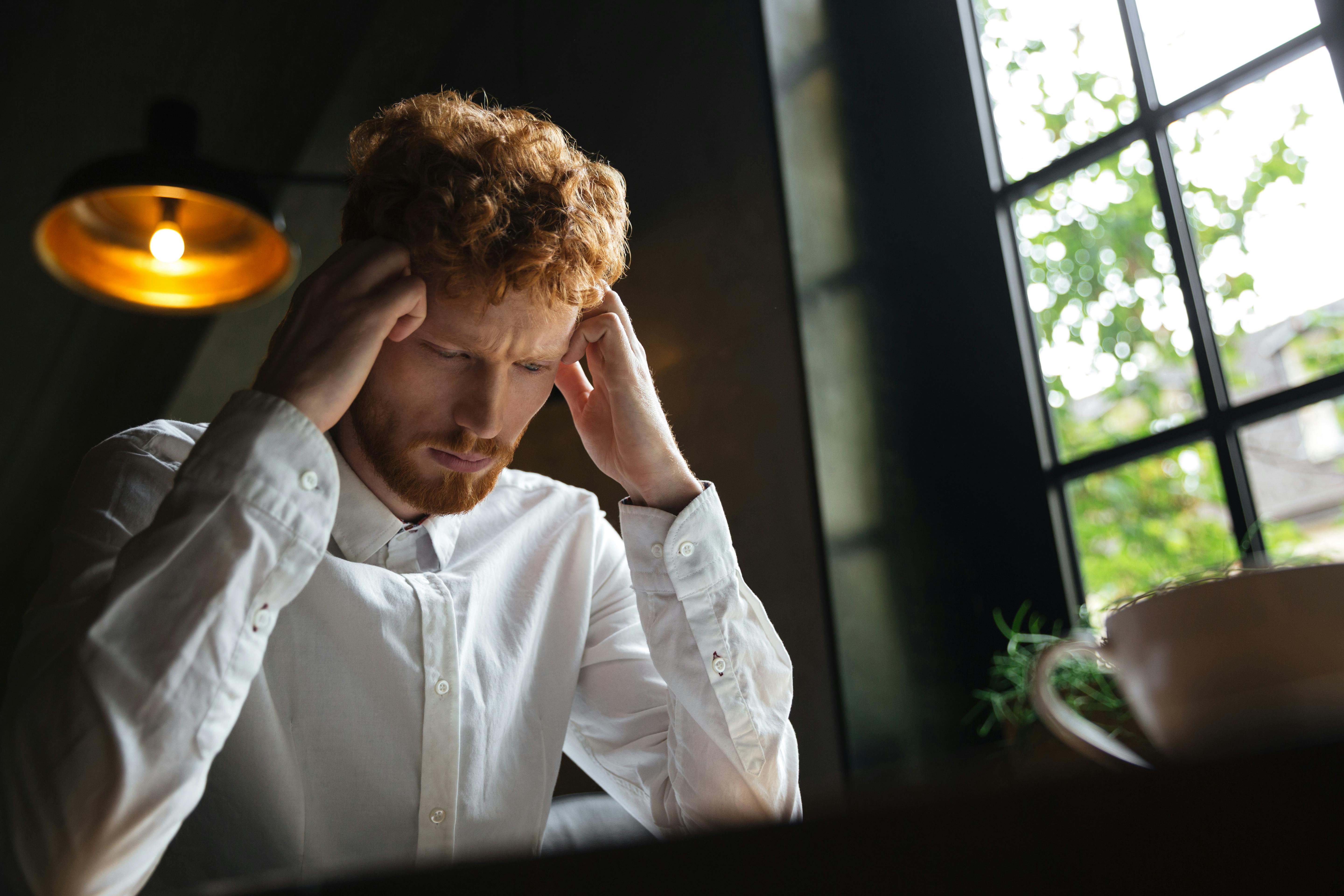 Close-up of redhead bearded man in white shirt looking stressed while sitting at a table.