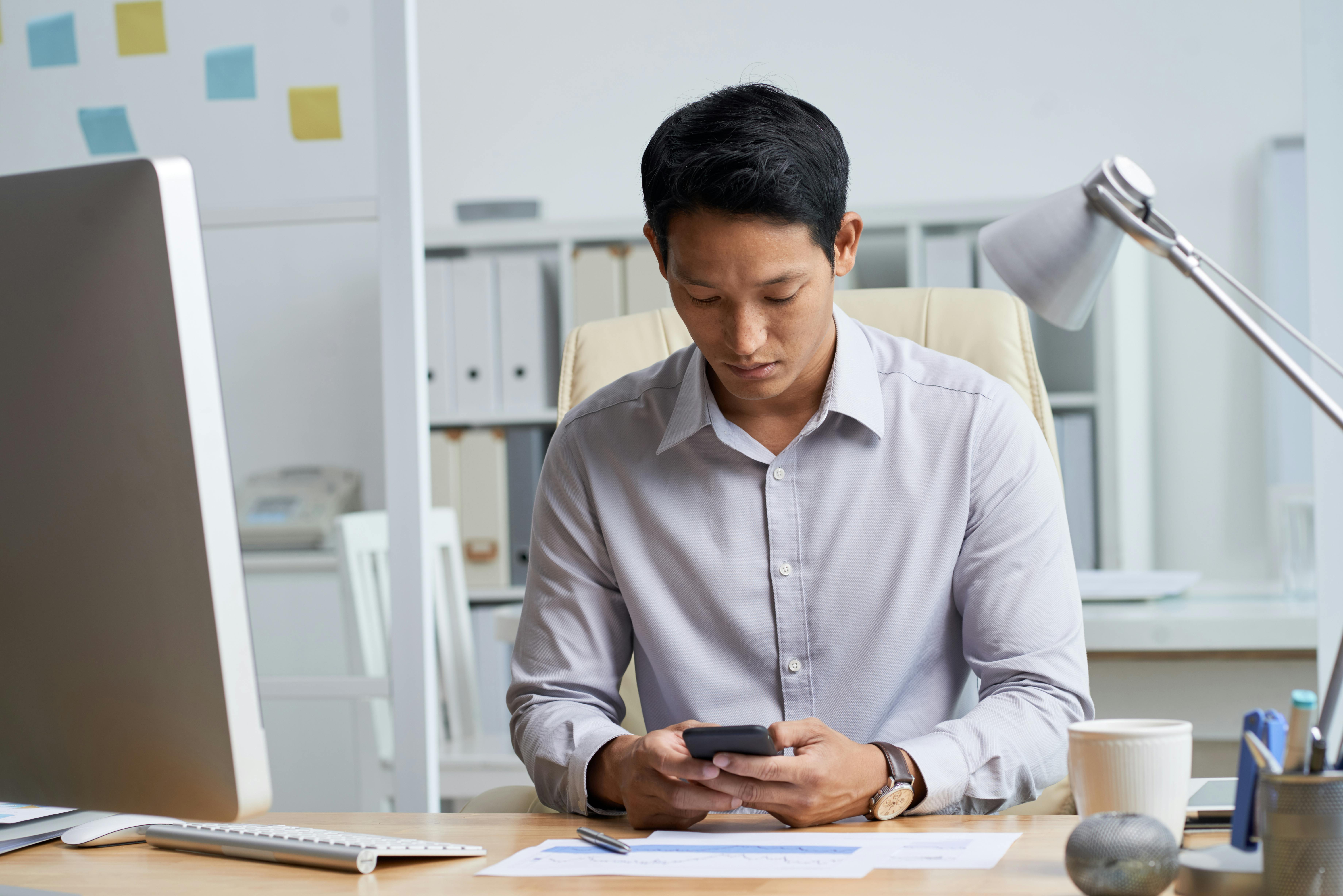 Asian man on cellphone can’t focus on one task while at desk in office
