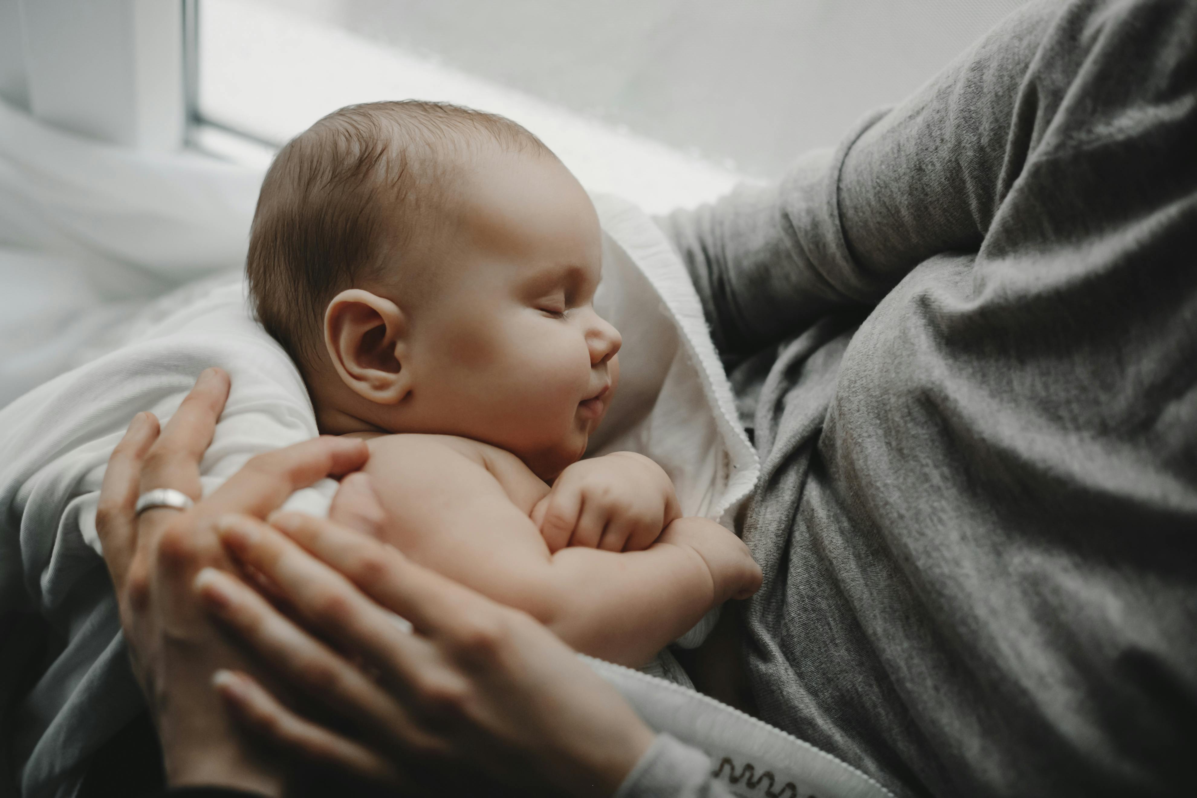 White newborn baby sleeping in white parent’s arms at home near a window