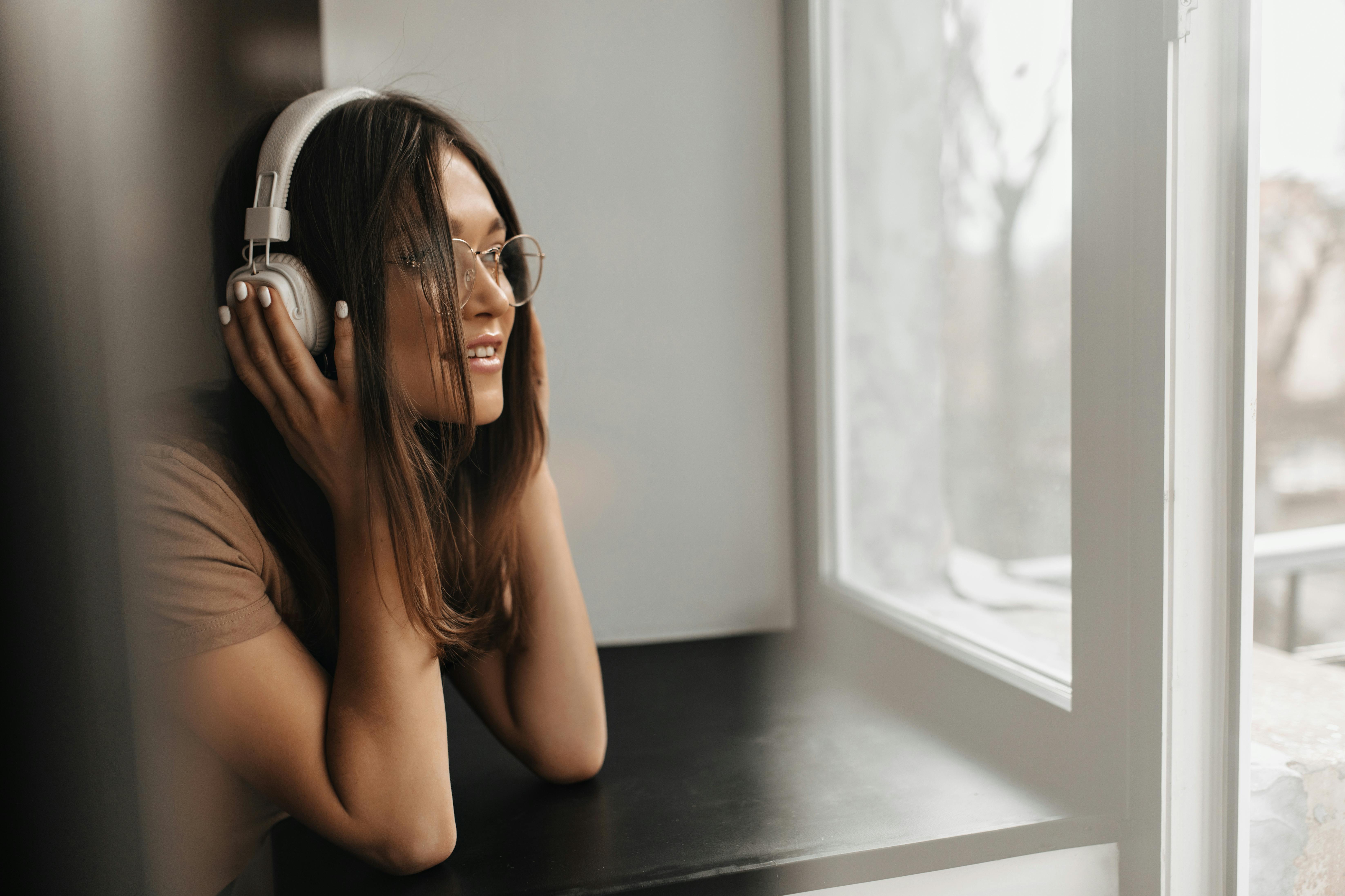 Young tan woman looks calm while listening to music and looking out a window. 