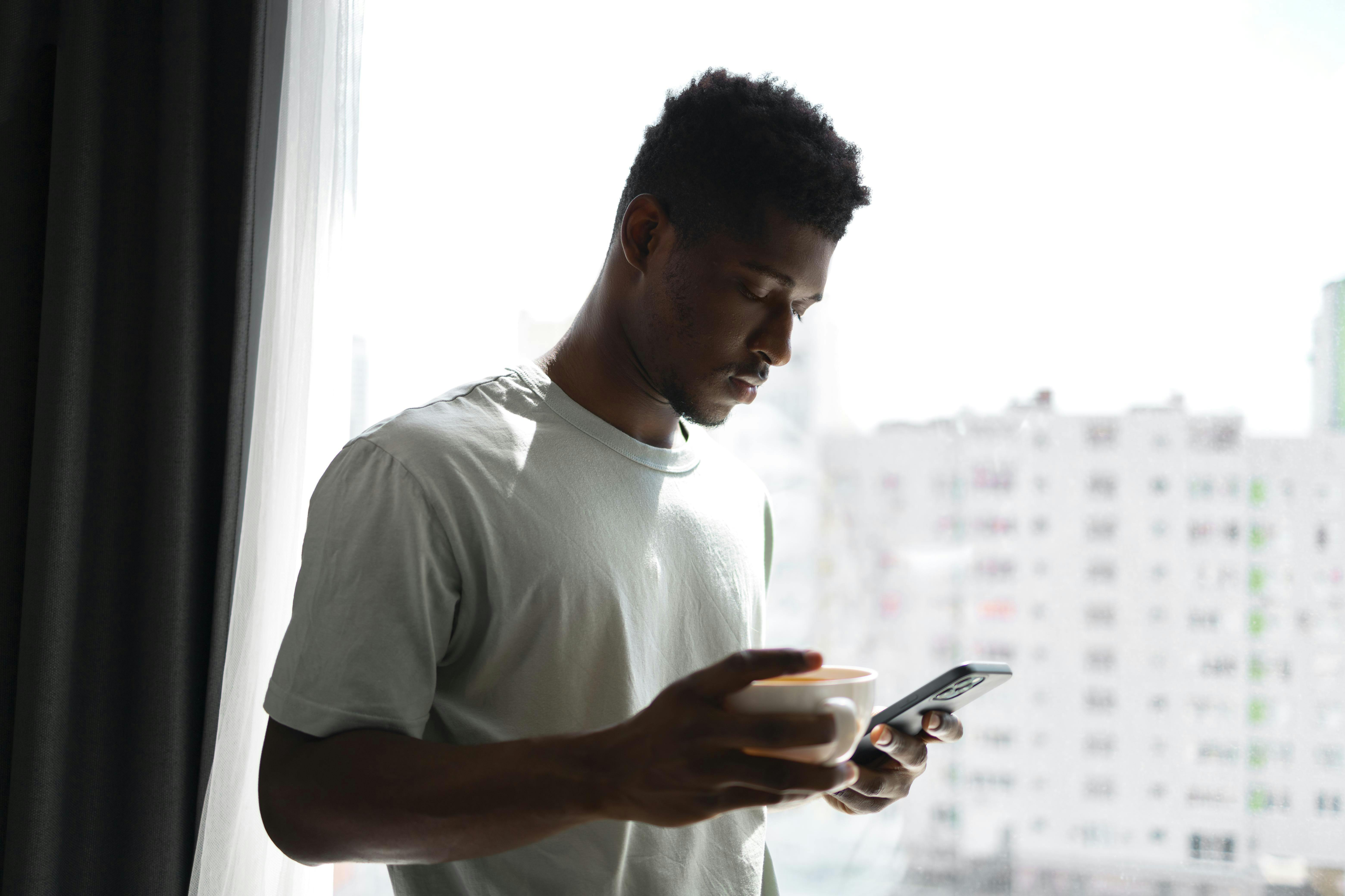 Black man uses phone in room while holding cup and looking focused and involved in task