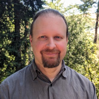 Cory Barr headshot. He wears a dark gray button-up shirt and stands in front of trees. He has dark hair, a mustache, and a goatee. He is smiling.