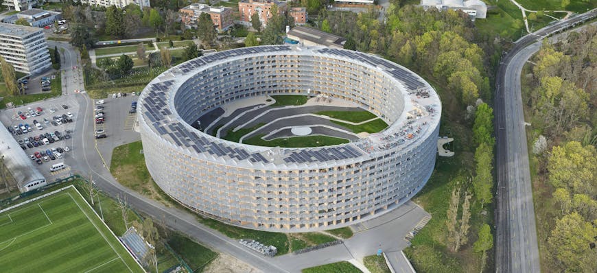 UHD 3D model of The Vortex in Lausanne, Switzerland, by Uzufly. Gray multistory cylindrical building with solar panels on top and roads and grass in the middle.
