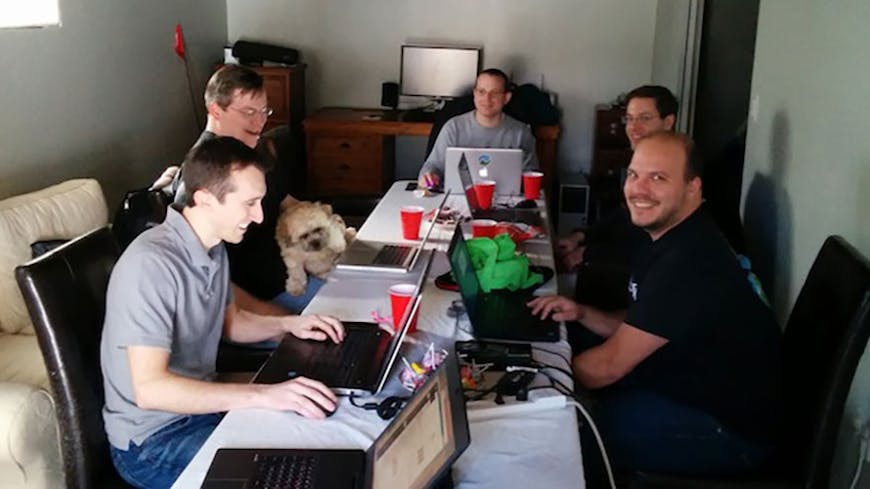 Five men sitting around a table each with a laptop. There is a dog on one person's lap. and everyone is smilling.