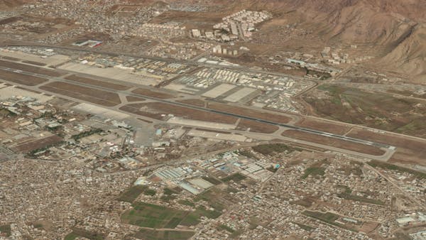 Maxar Precision3D Surface Model of Kabul International Airport. The airstrip runs diagonally from top left to bottom right. White planes are parked toward the middle of the image. The area is bounded on the top right by mountains. Fields and other buildings appear in the bottom half of the image.