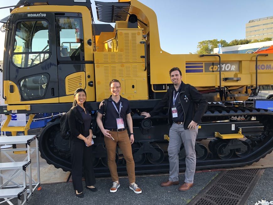 Cesium team members Mizuki Kobayashi Harris, Yonathan Anatolii Ast, and Ben Jewett pose in front of a Komatsu machine at the CSPI Expo.