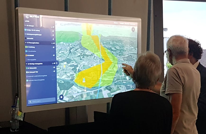 Gilytics' Pathfinder used in an interactive public engagement activity. A large screen in left-center displays Pathfinder. People in right-center touch the screen and examine the digital 3D model.