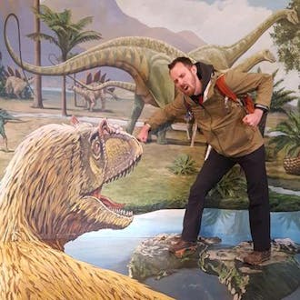 Mark Dane pretends his arm is being bitten by a model of a dinosaur.