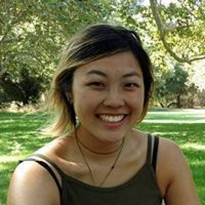 A woman in a green tank top stands in a park and smiles.