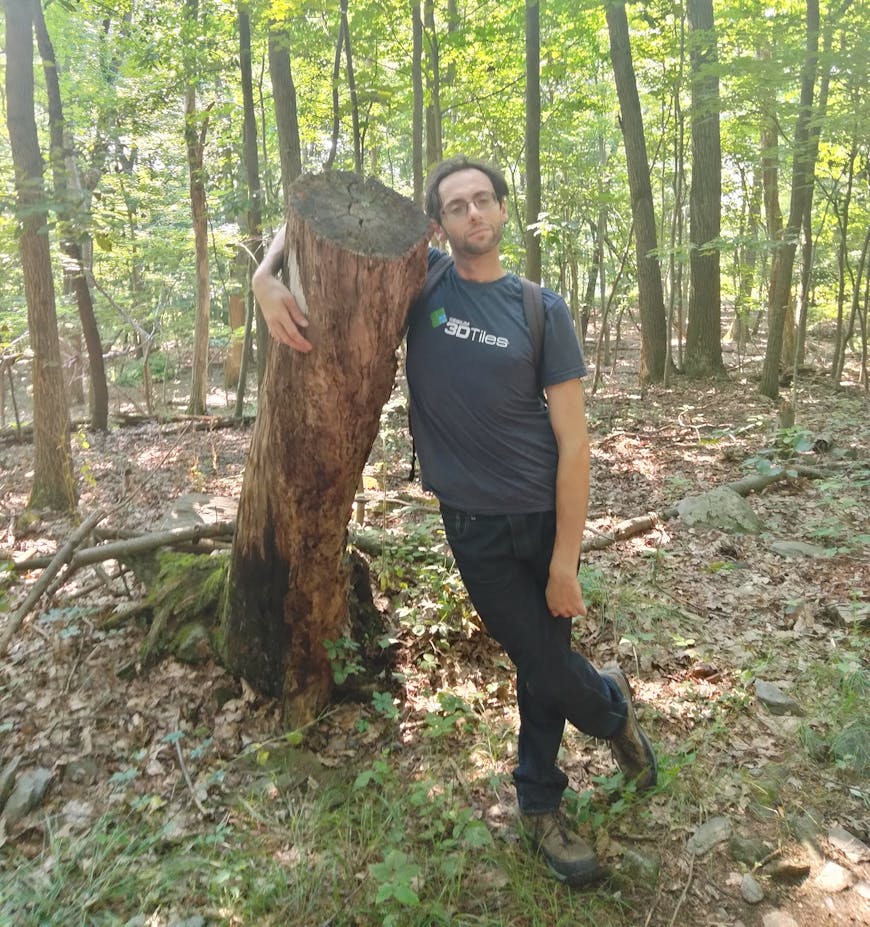 Daniel Krupka, wearing a 3D Tiles shirt, stands in the woods with his arm around a tall tree stump. 